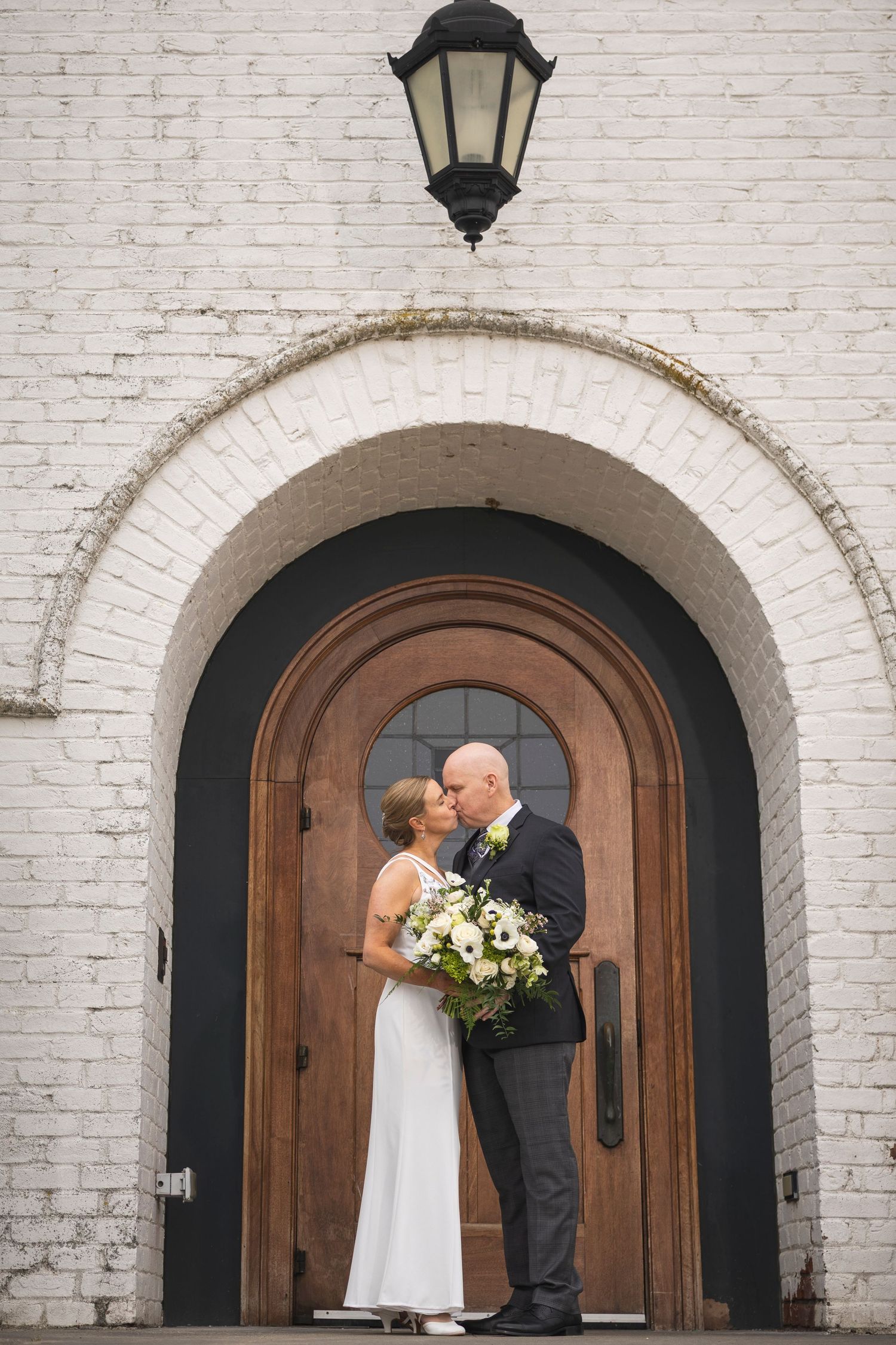 Wedding couple shares intimate moment against white brick wall with arched wooden door.