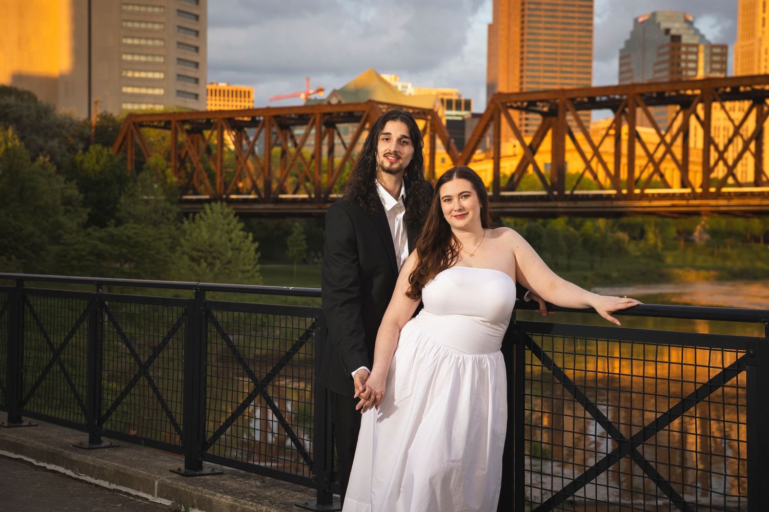 A couple poses on a bridge with a city skyline and railroad track in the background at sunset.