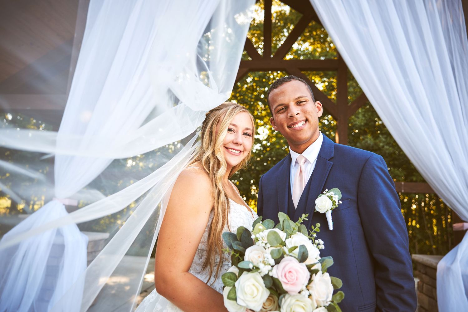 Wedding couple poses under elegant white drapes and floral decorations in an outdoor ceremony setting.