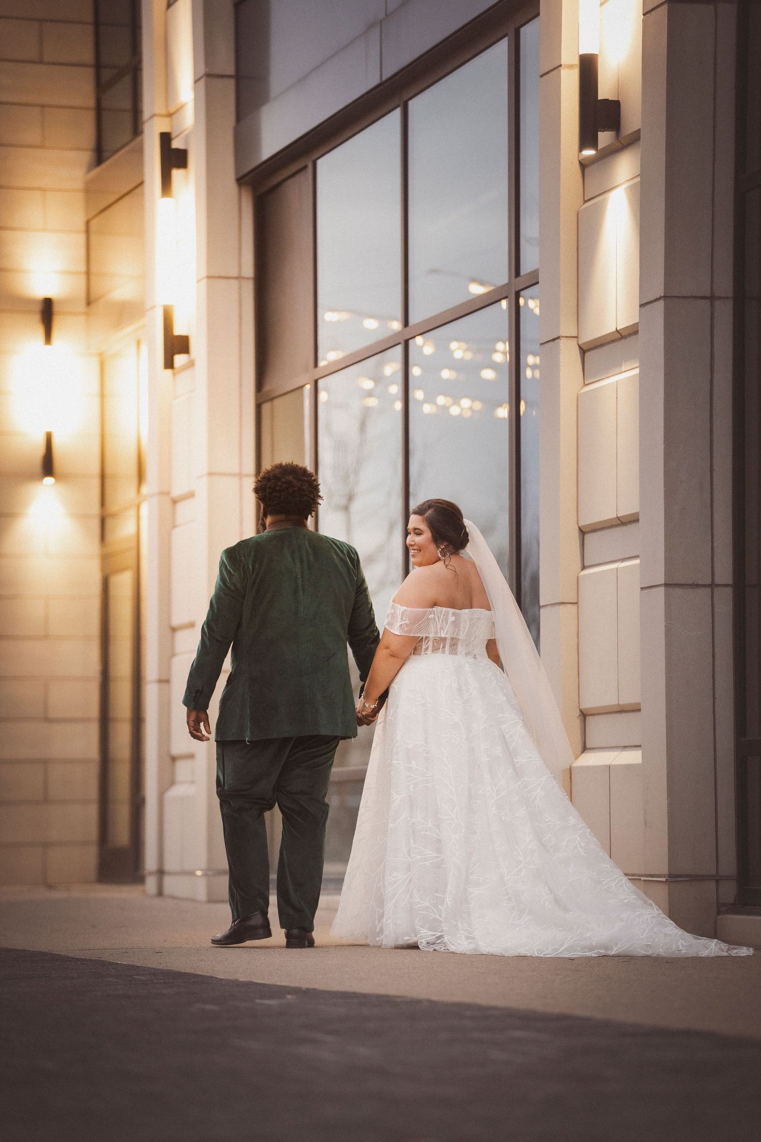 Elegant bride in white gown and groom in dark suit walk along modern building exterior.