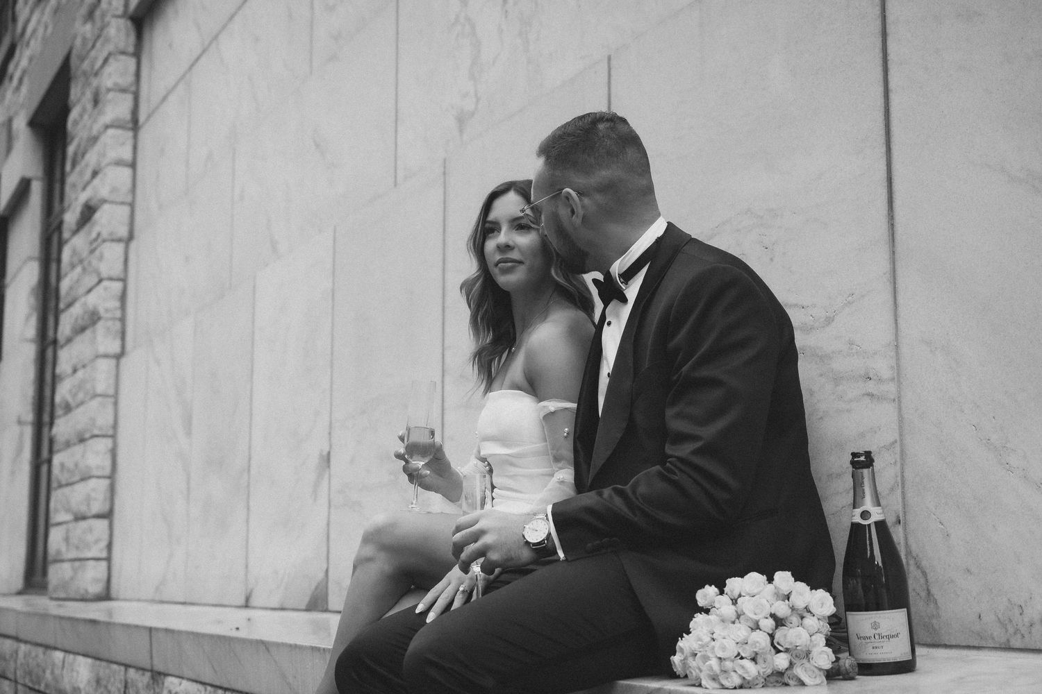 A romantic black and white wedding photo of a couple sitting together with champagne and flowers.