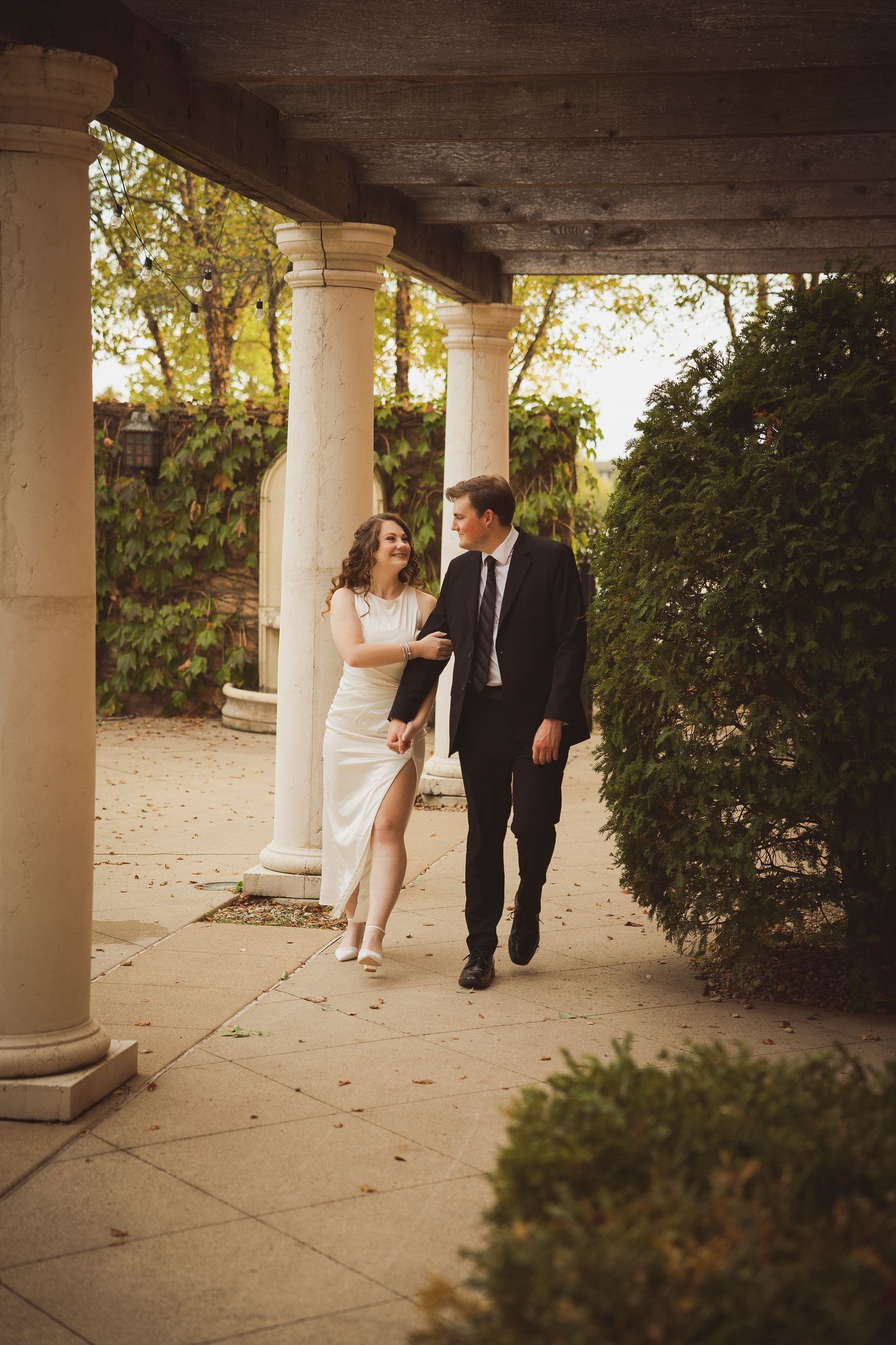 A couple strolls beneath white columns and archways during sunset.