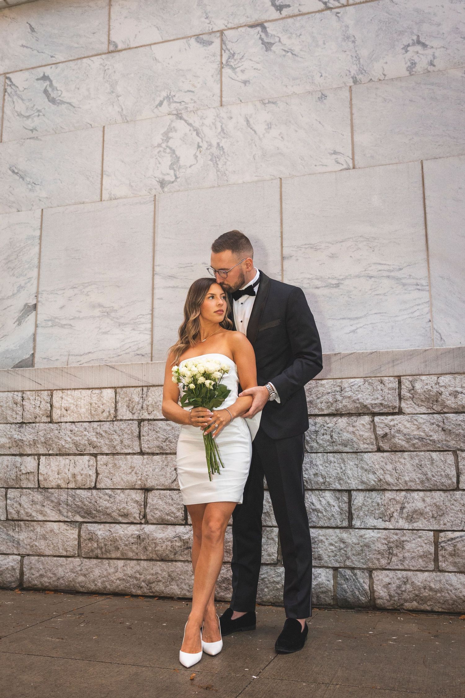 A elegant couple in formal attire posing against a marble wall with a white bouquet.