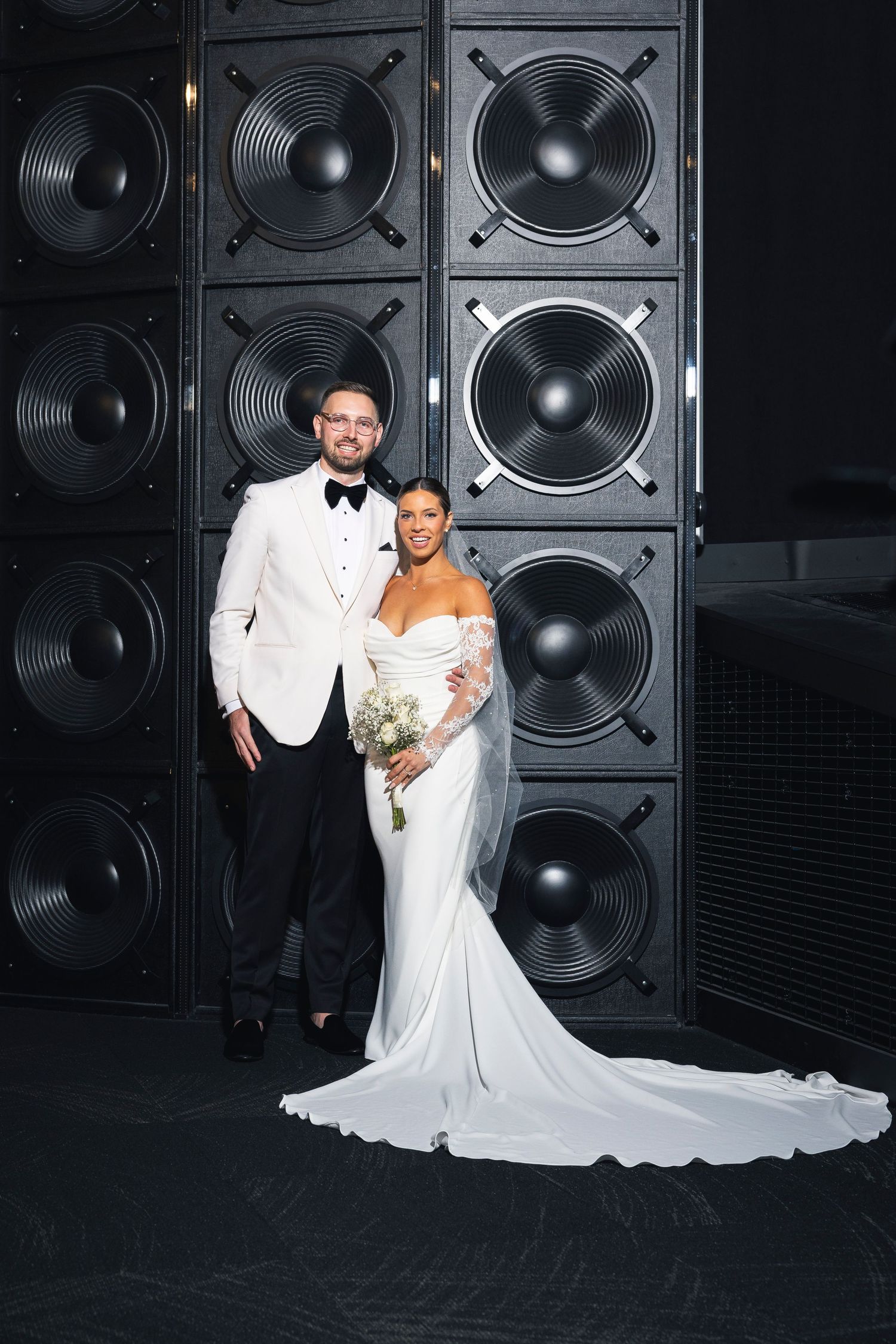 Newlyweds pose for photos in front of a wall of black speakers at their reception venue.