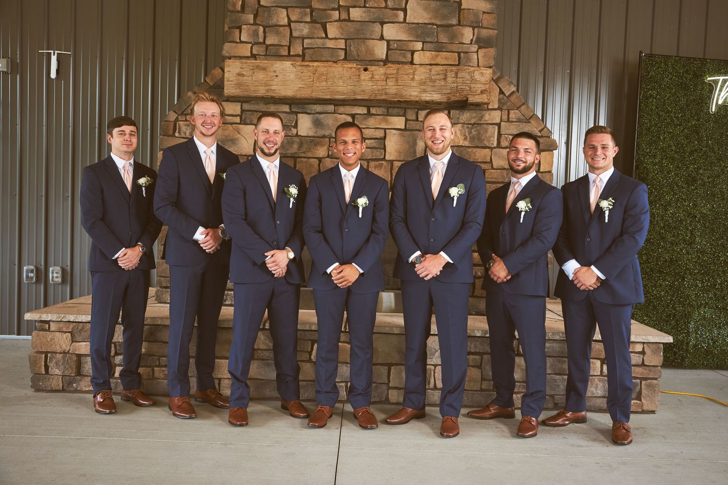 Groomsmen in navy suits and brown shoes posed sitting on stone wall.