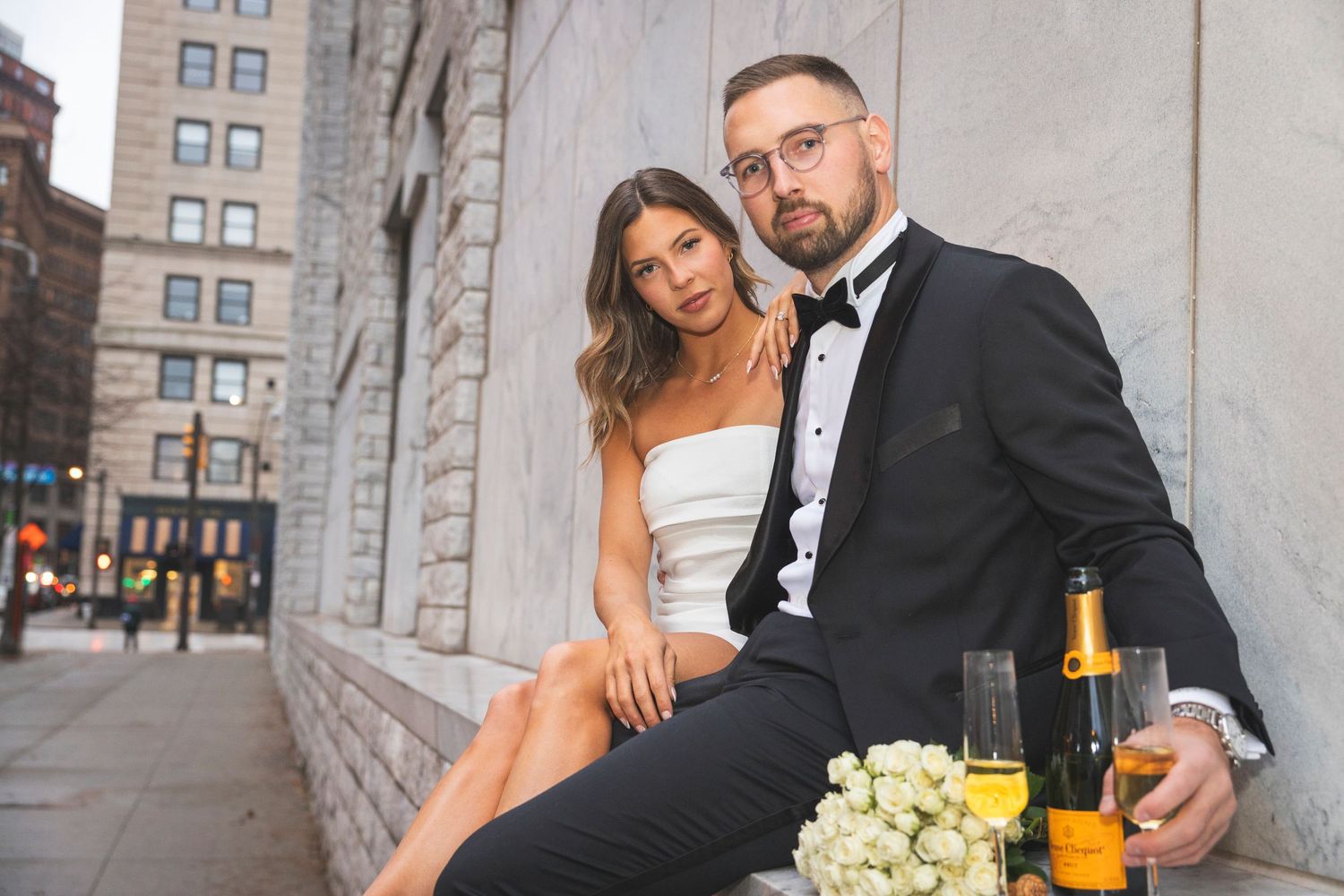 Wedding couple poses near stone building with urban backdrop.