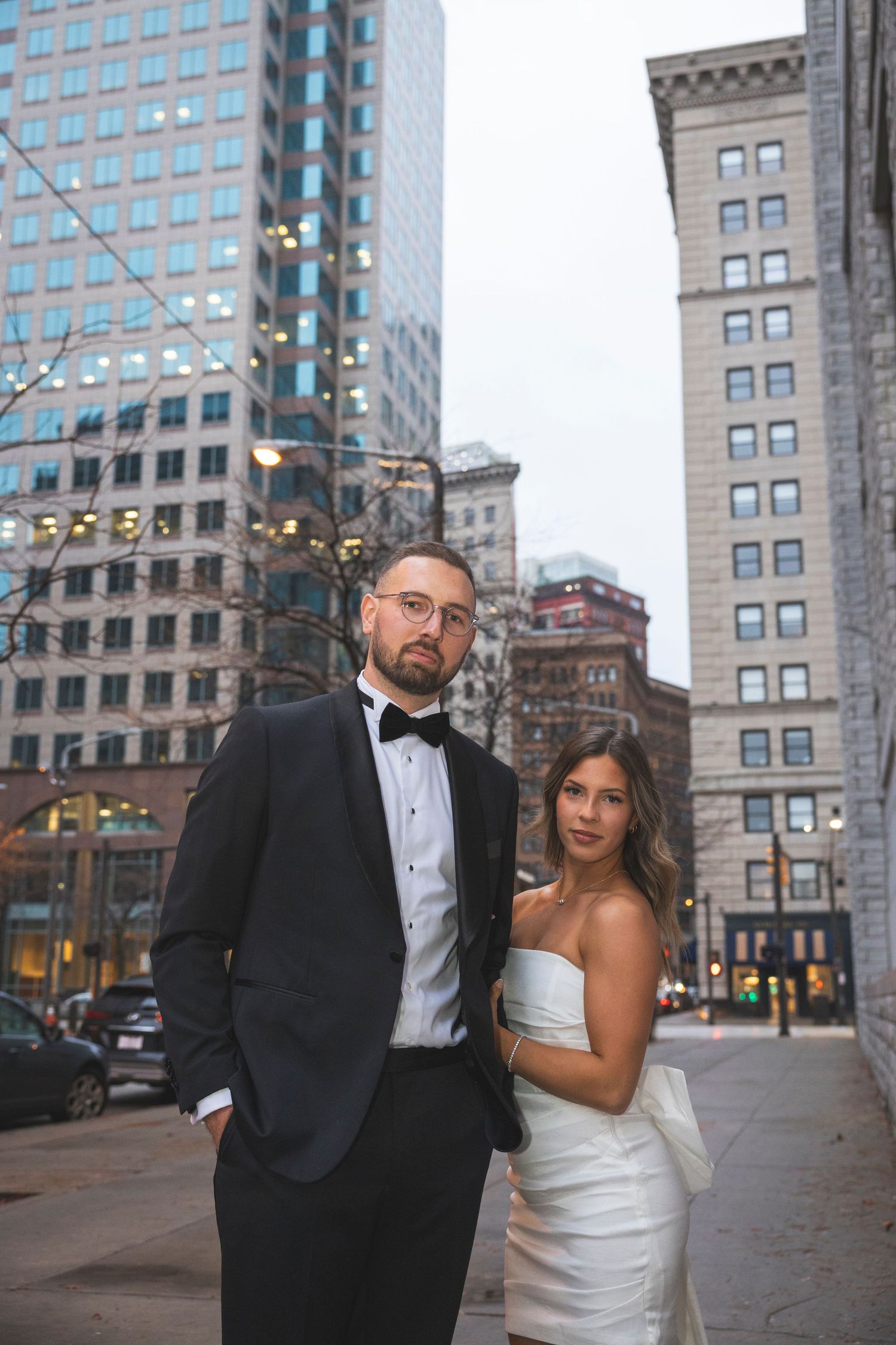 A formal couple pose for wedding photos in front of city buildings at dusk.