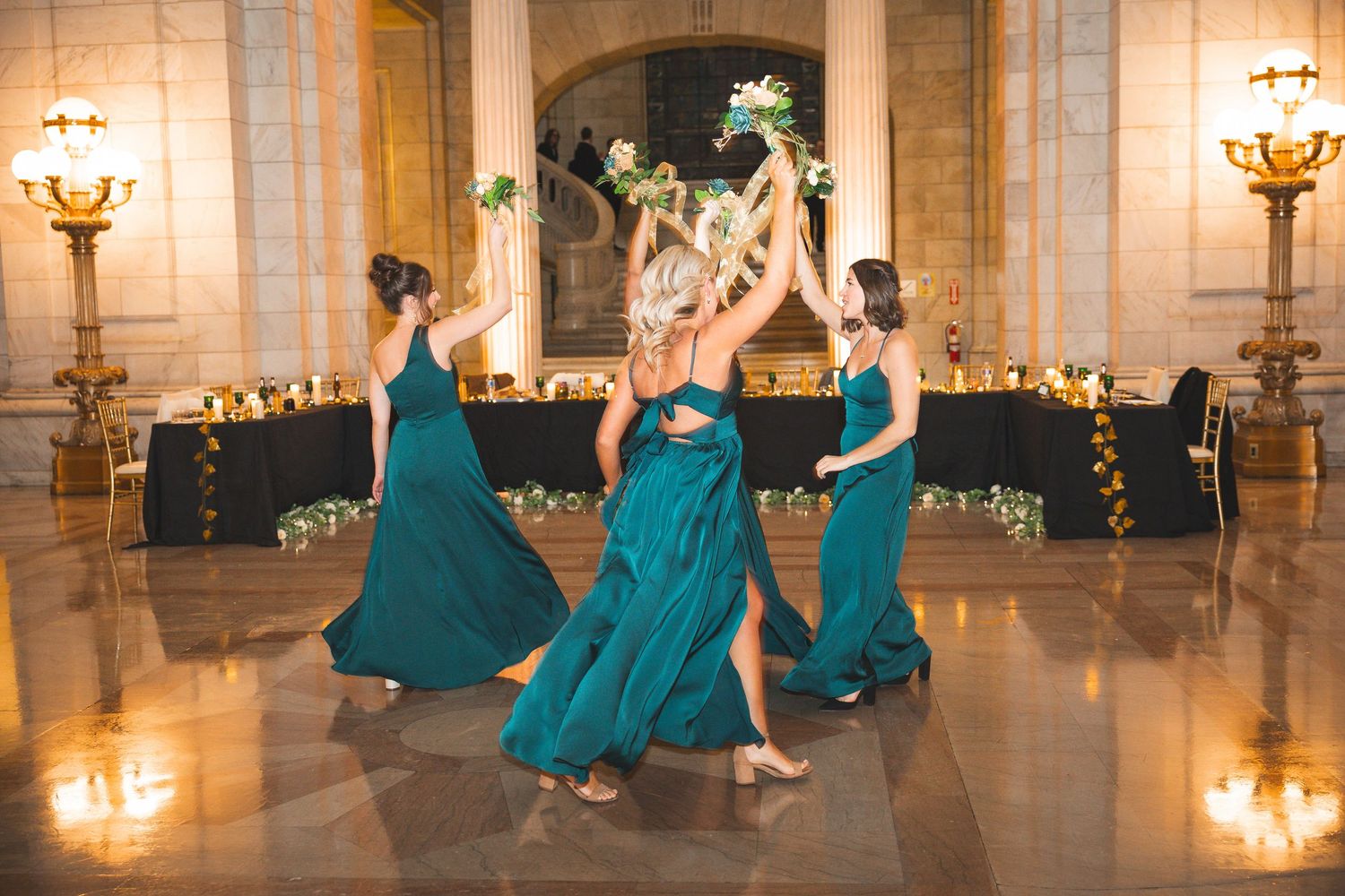 Bridesmaids in elegant teal dresses dance together in a grand ballroom with ornate lighting and architecture.