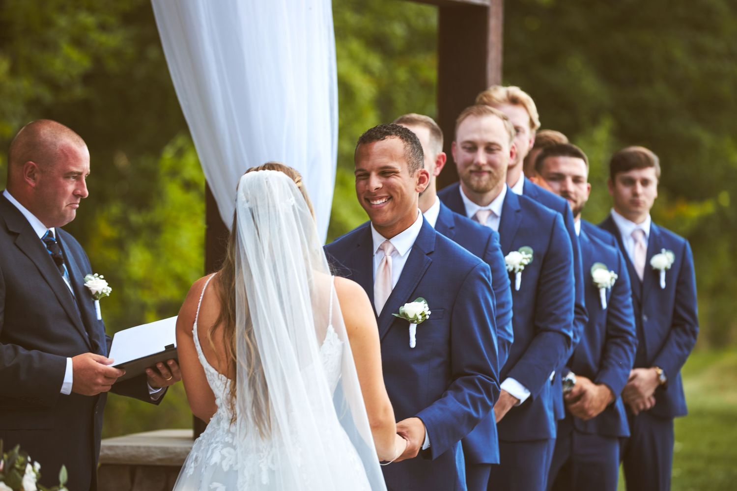 Wedding party members in navy blue suits stand together during an outdoor ceremony.
