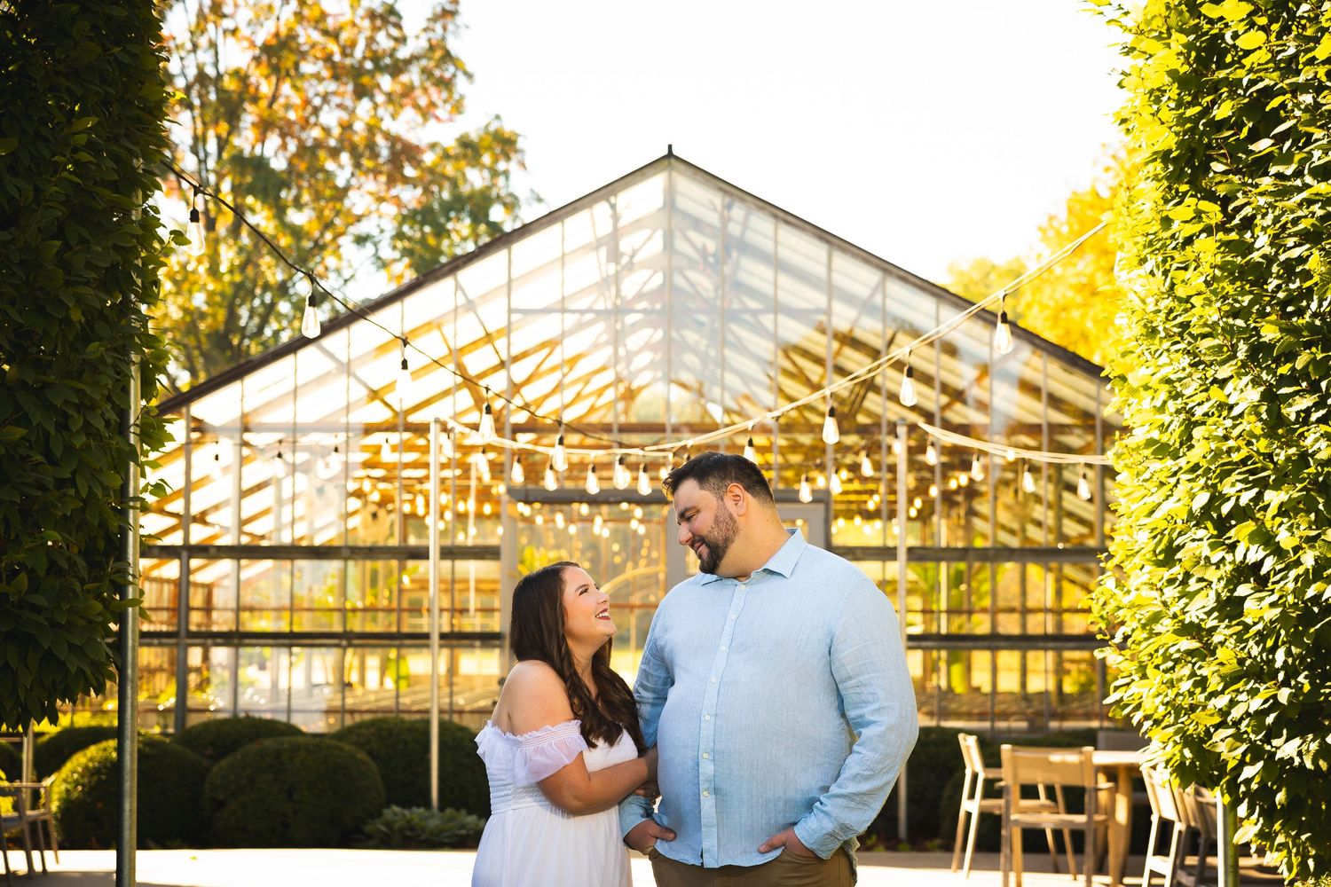 Two people share a moment in front of a sunlit greenhouse with glass panels and lush greenery.