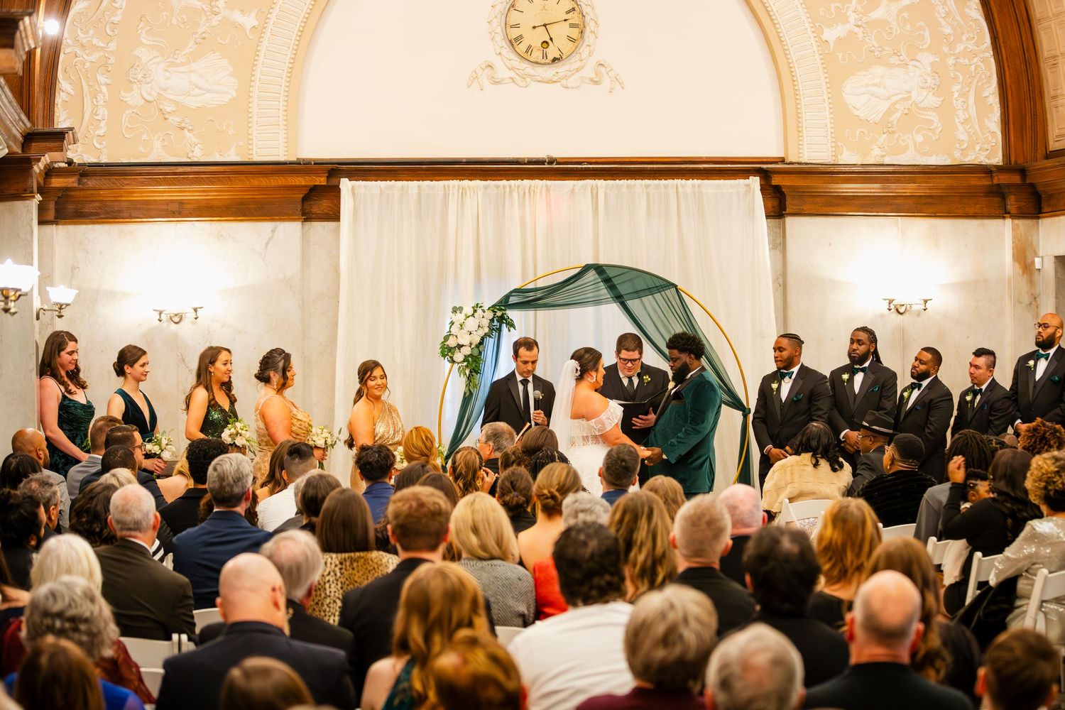 Wide view of a wedding ceremony with guests seated in rows under an ornate arched ceiling.
