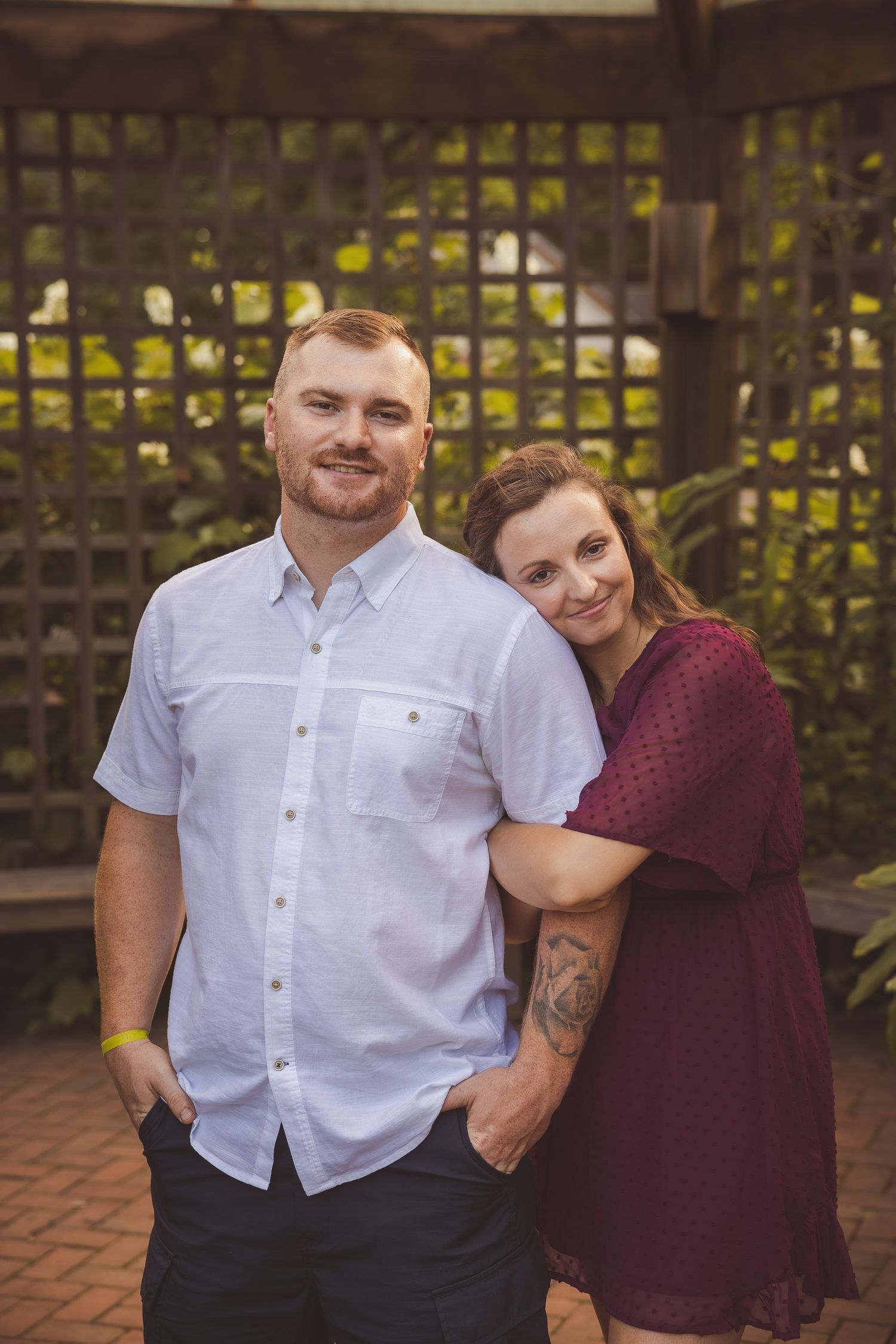 A couple in burgundy and white clothing stands together in front of a wooden lattice garden wall.