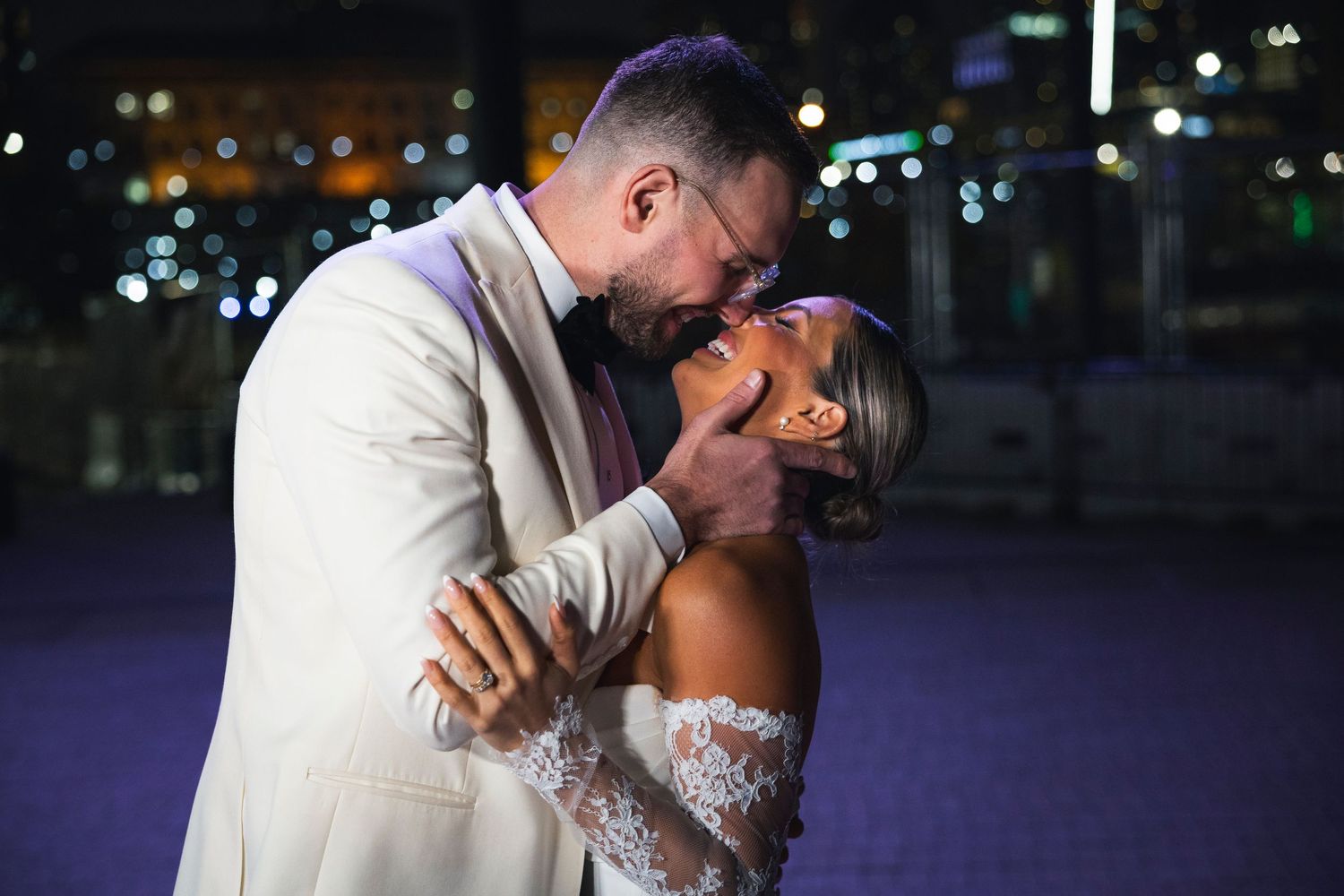 Intimate moment captured between couple during nighttime wedding photoshoot with city lights in background.
