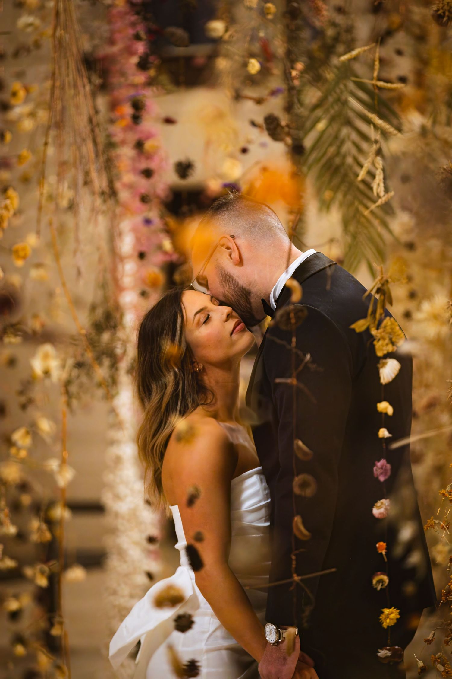Romantic wedding moment captured under hanging floral decorations with golden lighting.