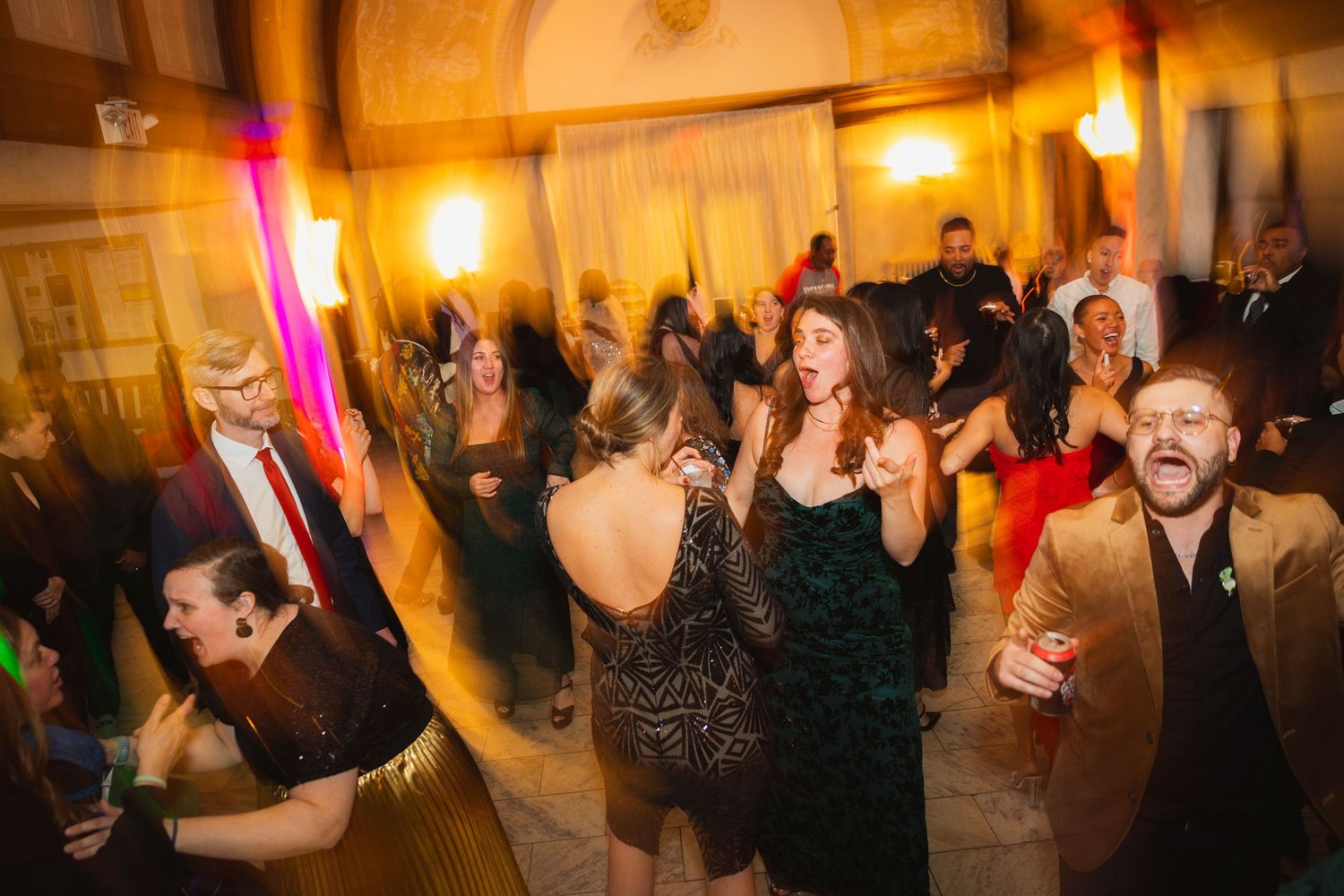Wedding guests dance and celebrate on a dimly lit dance floor with colorful party lights.