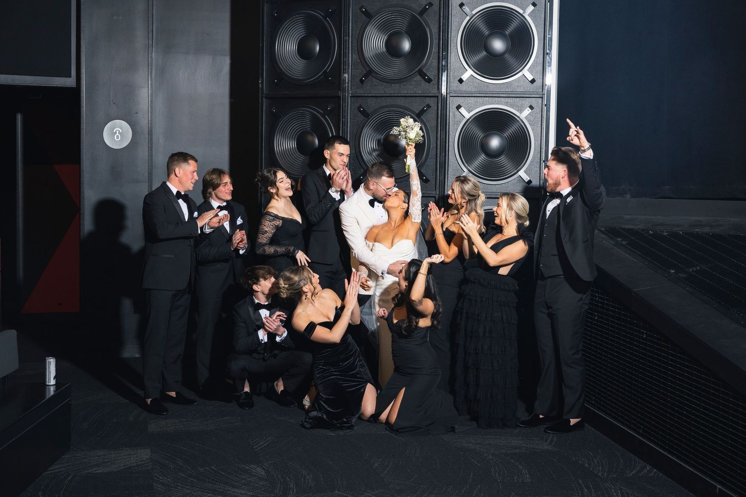 Wedding party gathers for photos in front of large speaker wall in dimly lit venue.