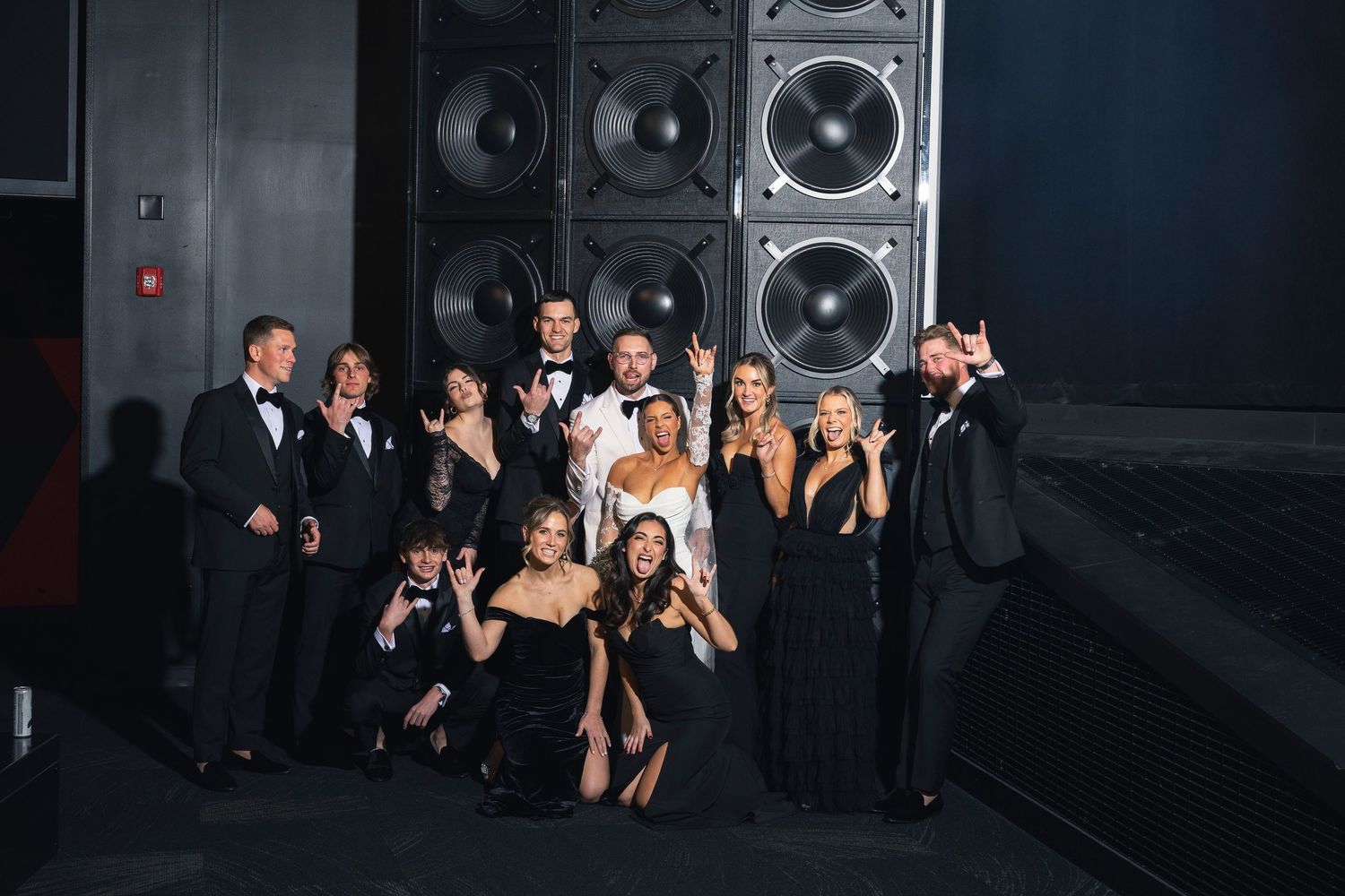 Wedding party members pose together in formal black attire against dark industrial backdrop.