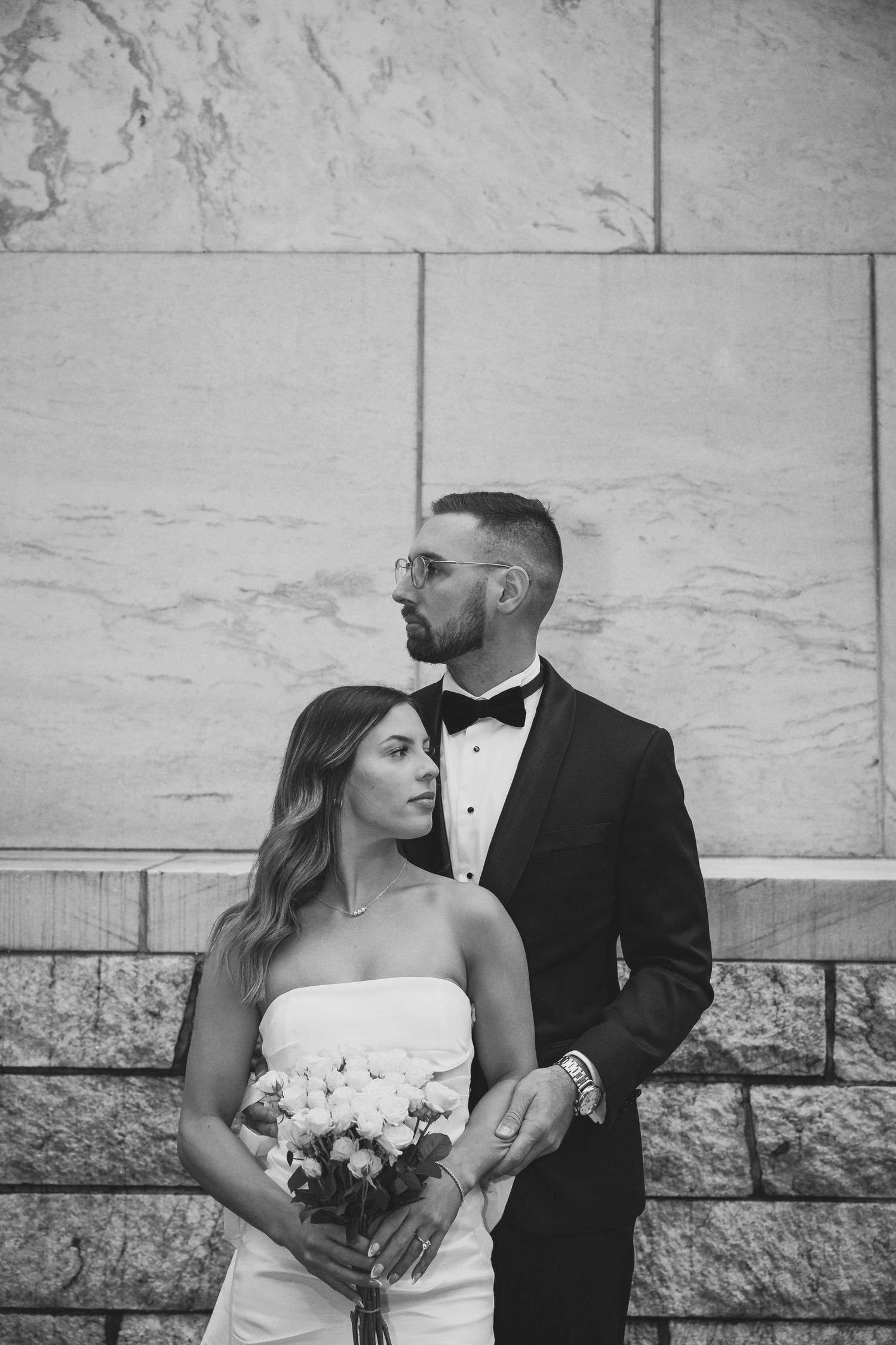 A black and white portrait of a couple in wedding attire against a stone wall.
