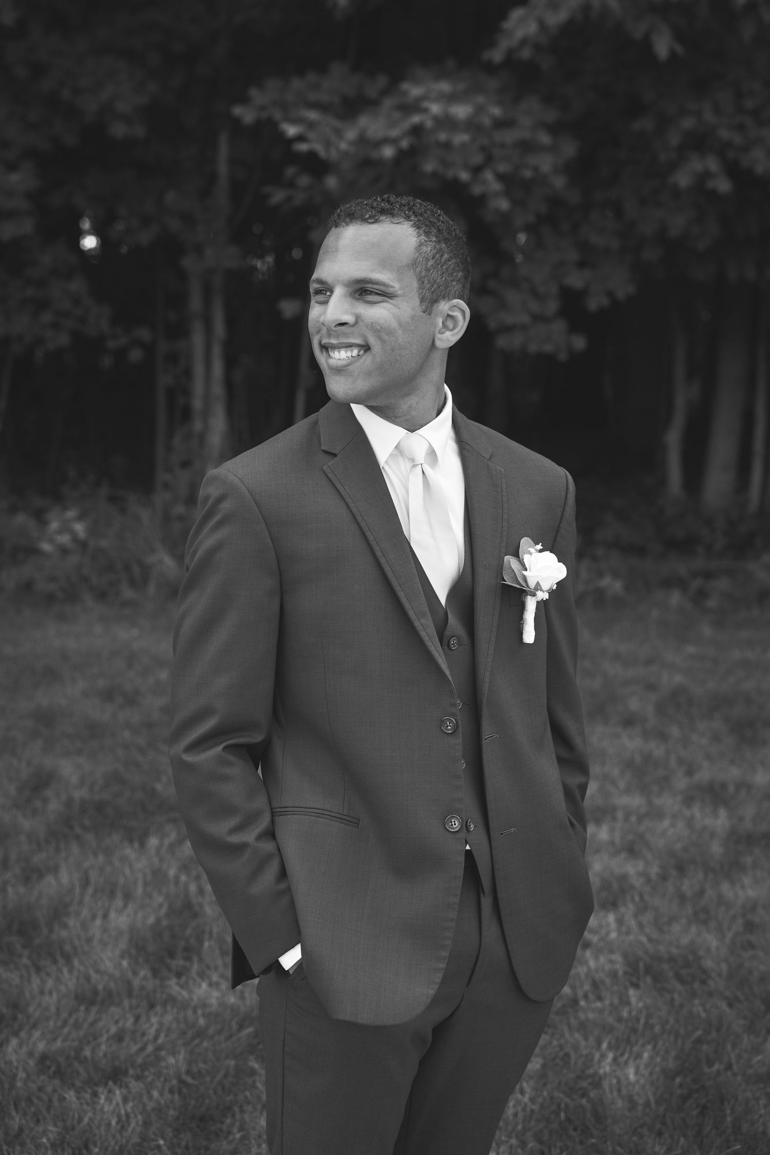 Black and white portrait of groomsman in formal suit with boutonniere smiling outdoors.