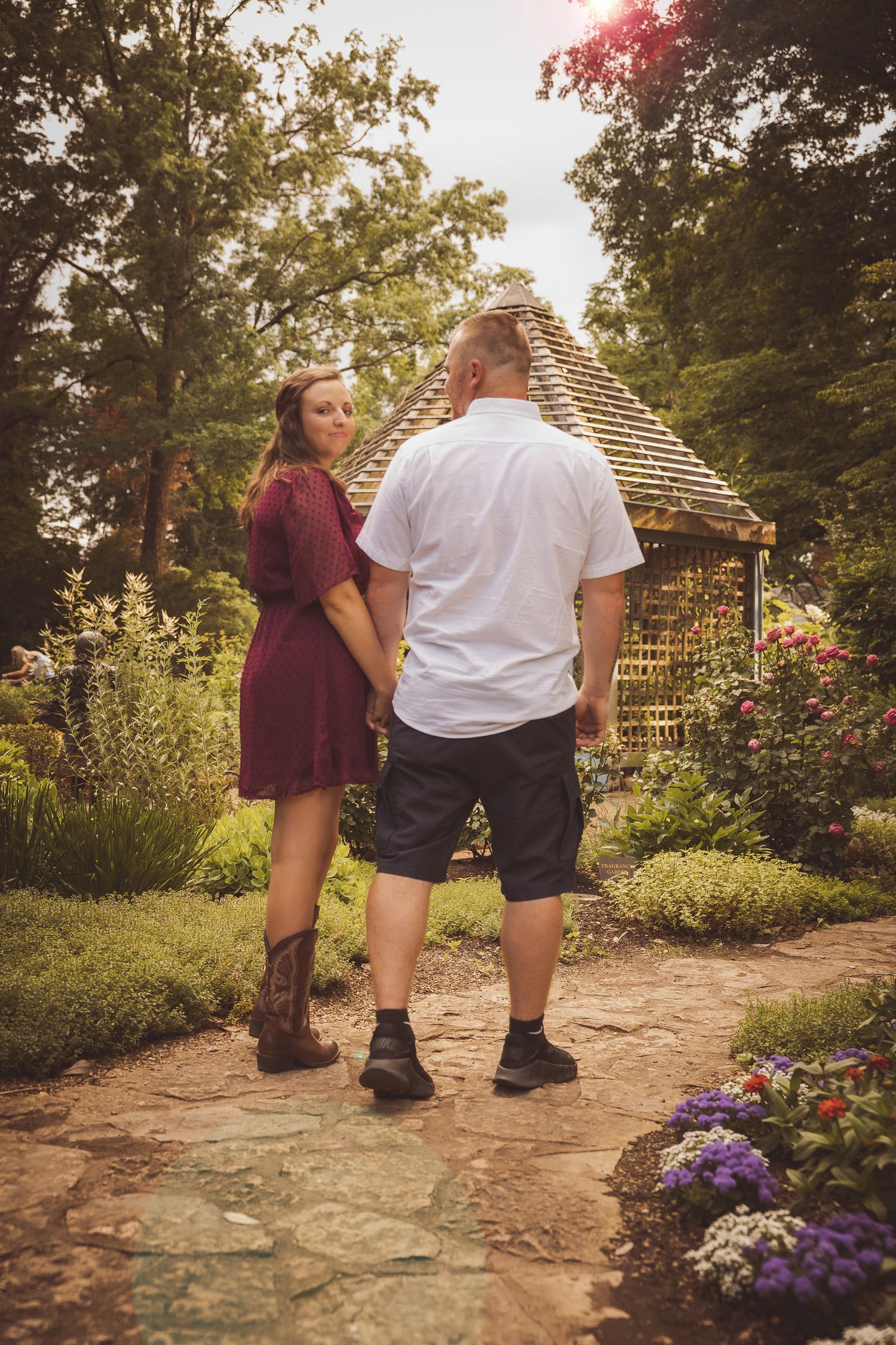 A couple walking together on a garden path with purple flowers and greenery on a sunny day.