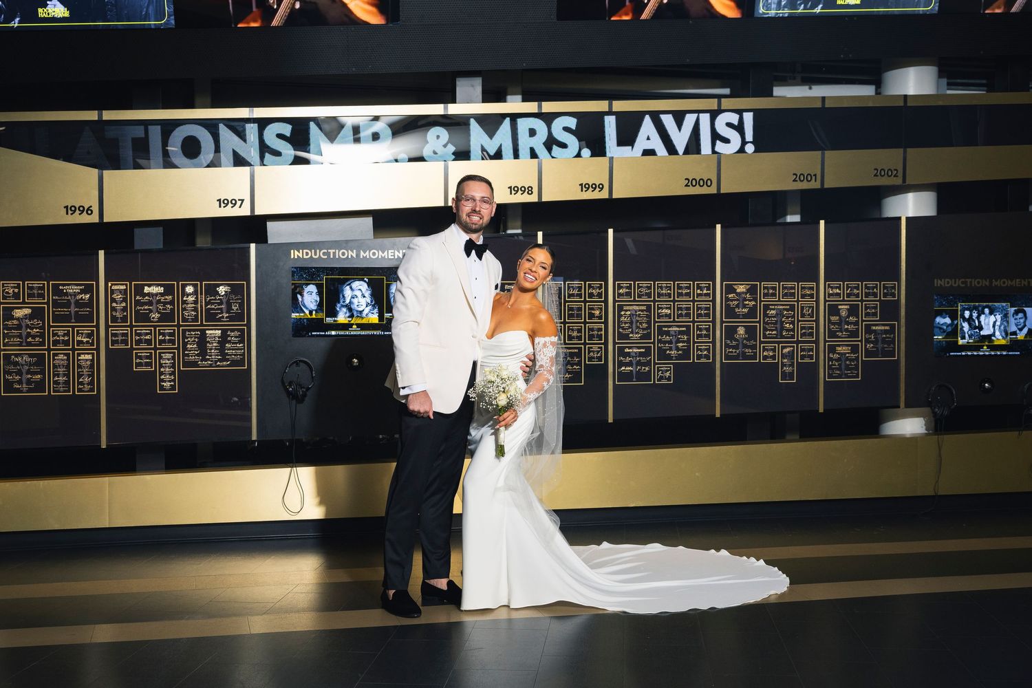 Wedding couple poses under 'MR & MRS LAVIS' sign in modern venue interior.