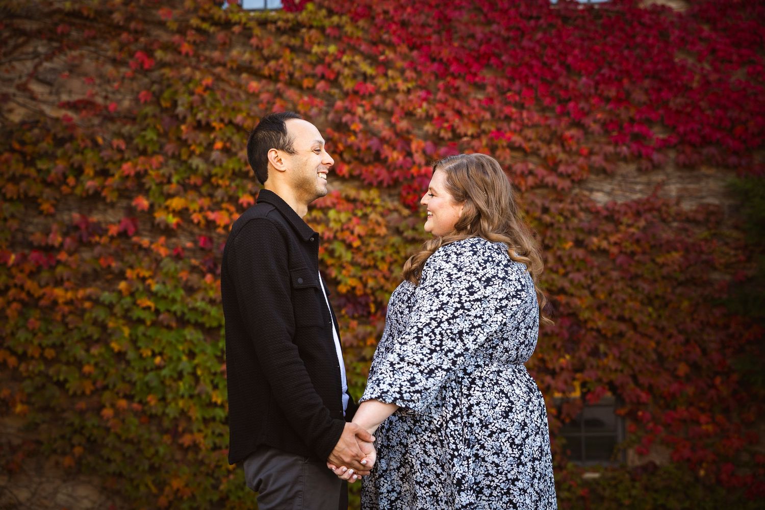 Two people share a romantic moment in front of a vibrant red autumn vine-covered wall.