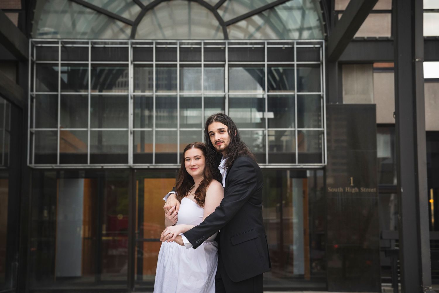 A couple poses under a modern glass canopy entrance during a formal photoshoot.