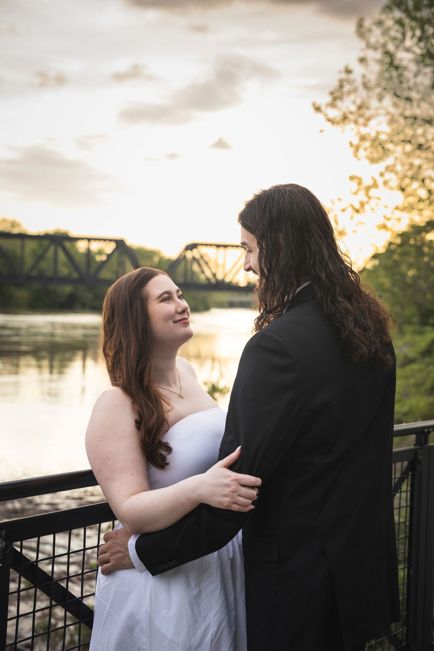 A couple embraces at sunset near a riverfront with a bridge visible in the background.