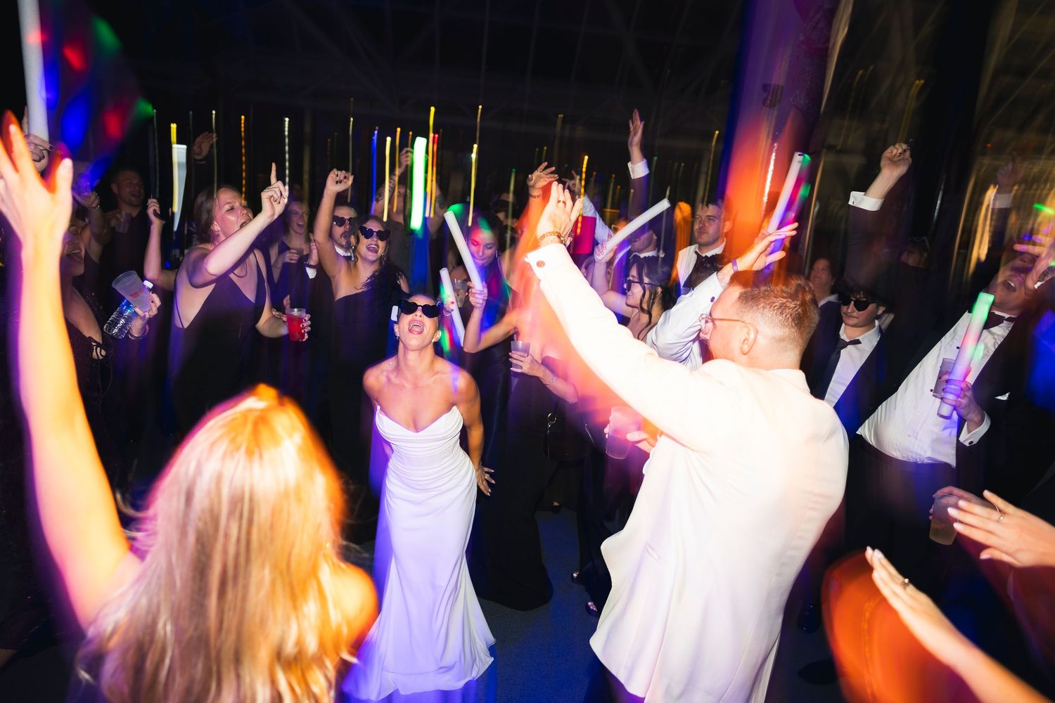 Wedding celebration with crowd dancing and raising hands under colorful lights.