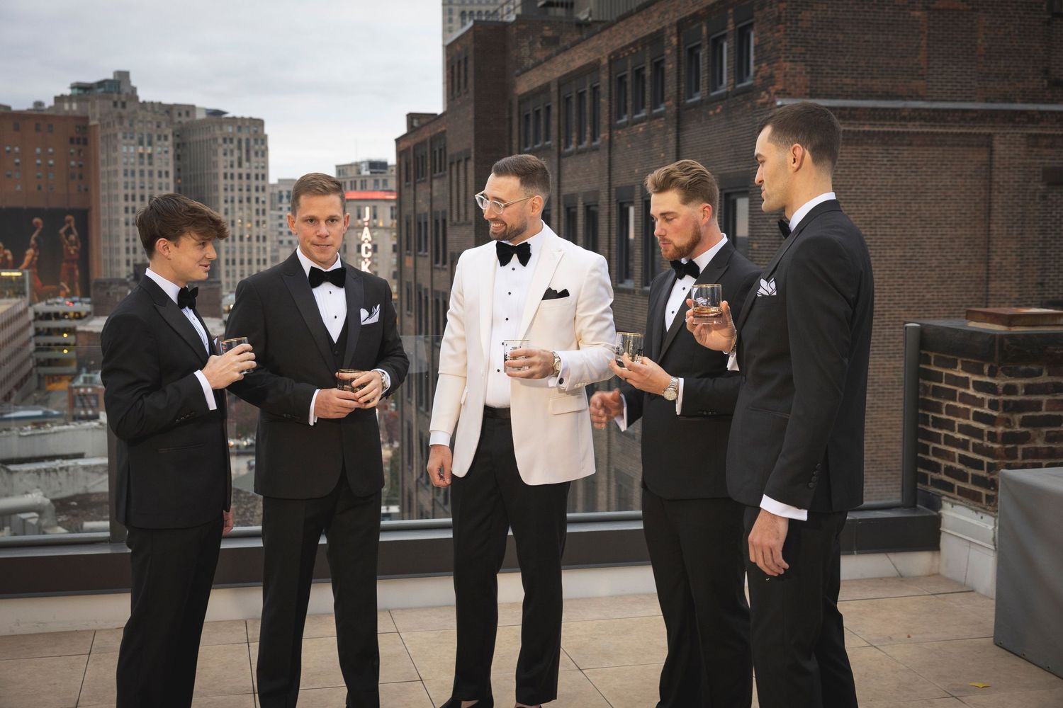 Wedding party members in tuxedos gather on a rooftop overlooking a city backdrop.