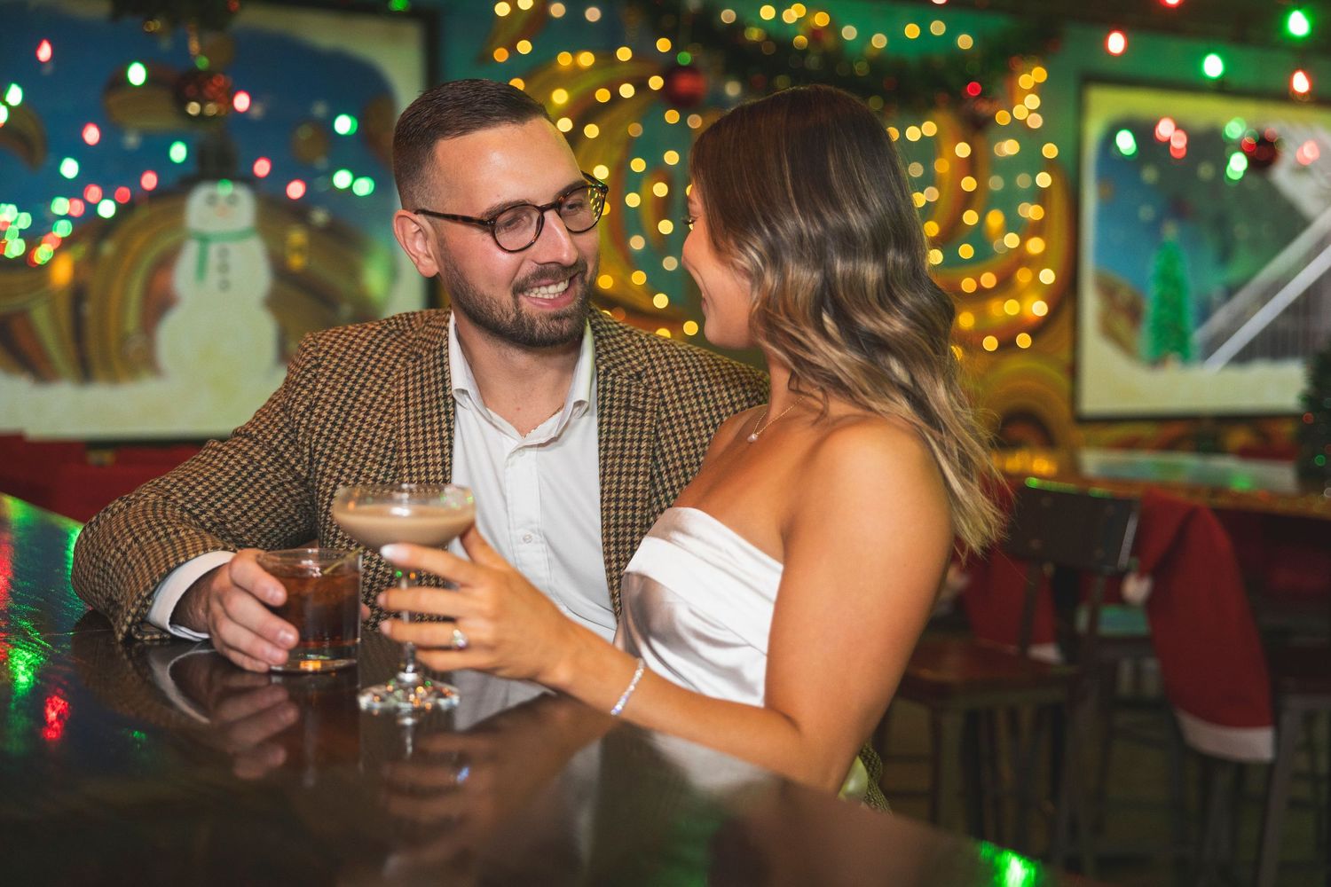 A couple shares an intimate moment at a bar decorated with festive holiday lights and decor.