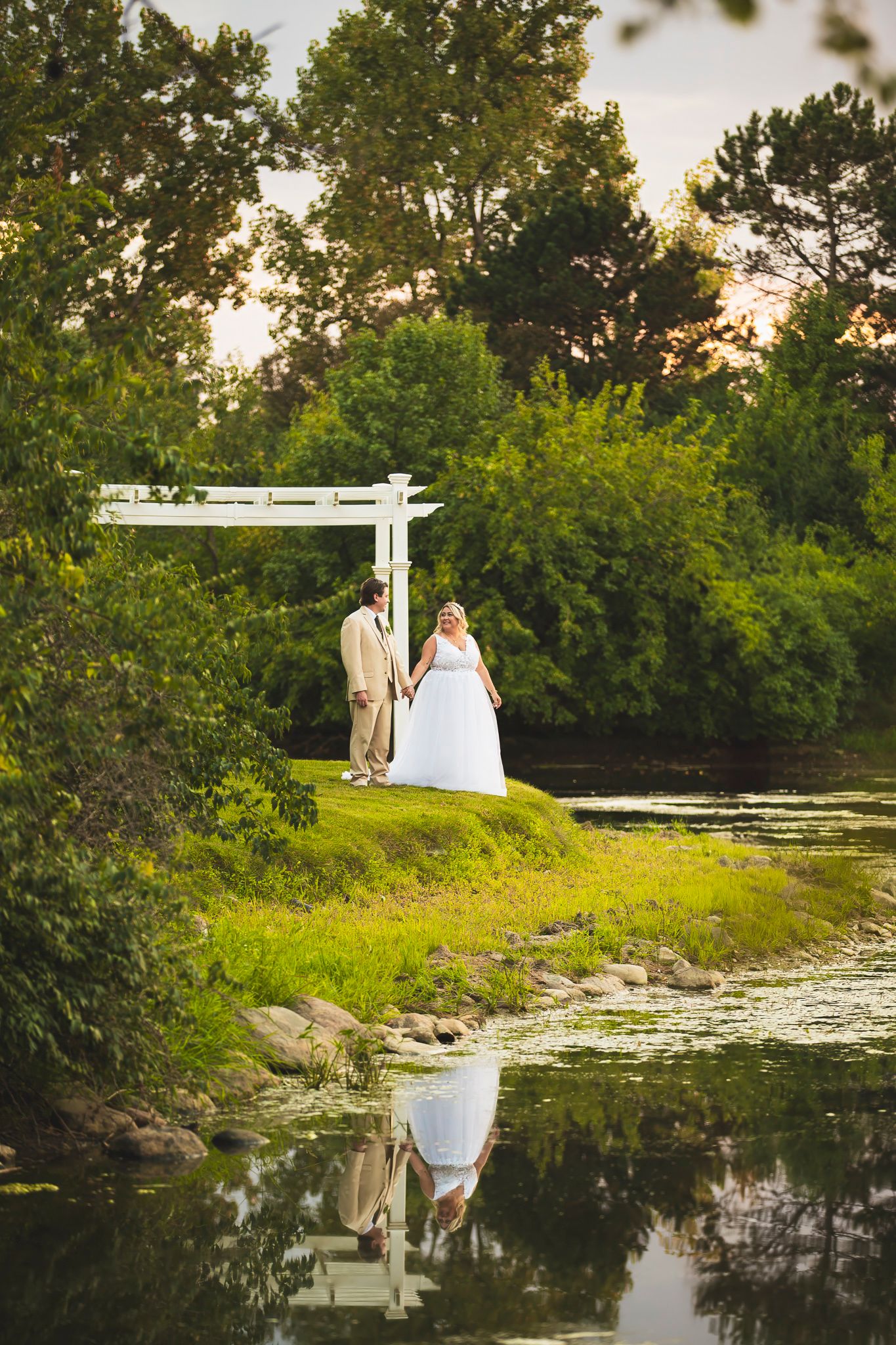 A scenic wedding ceremony setup near a tranquil pond with white decorations and lush greenery.