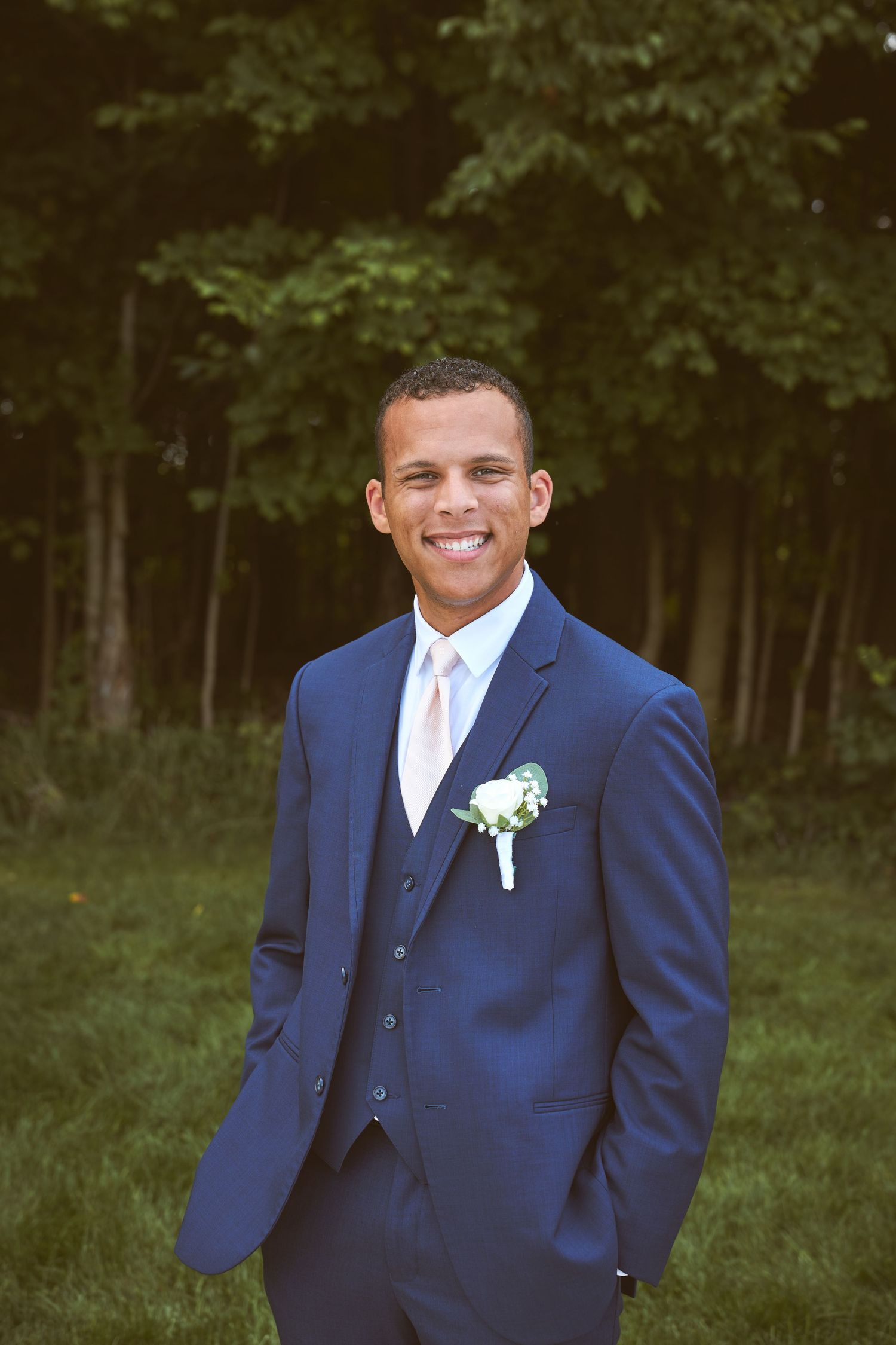 Groomsman in navy three-piece suit with white tie and boutonniere poses outdoors.
