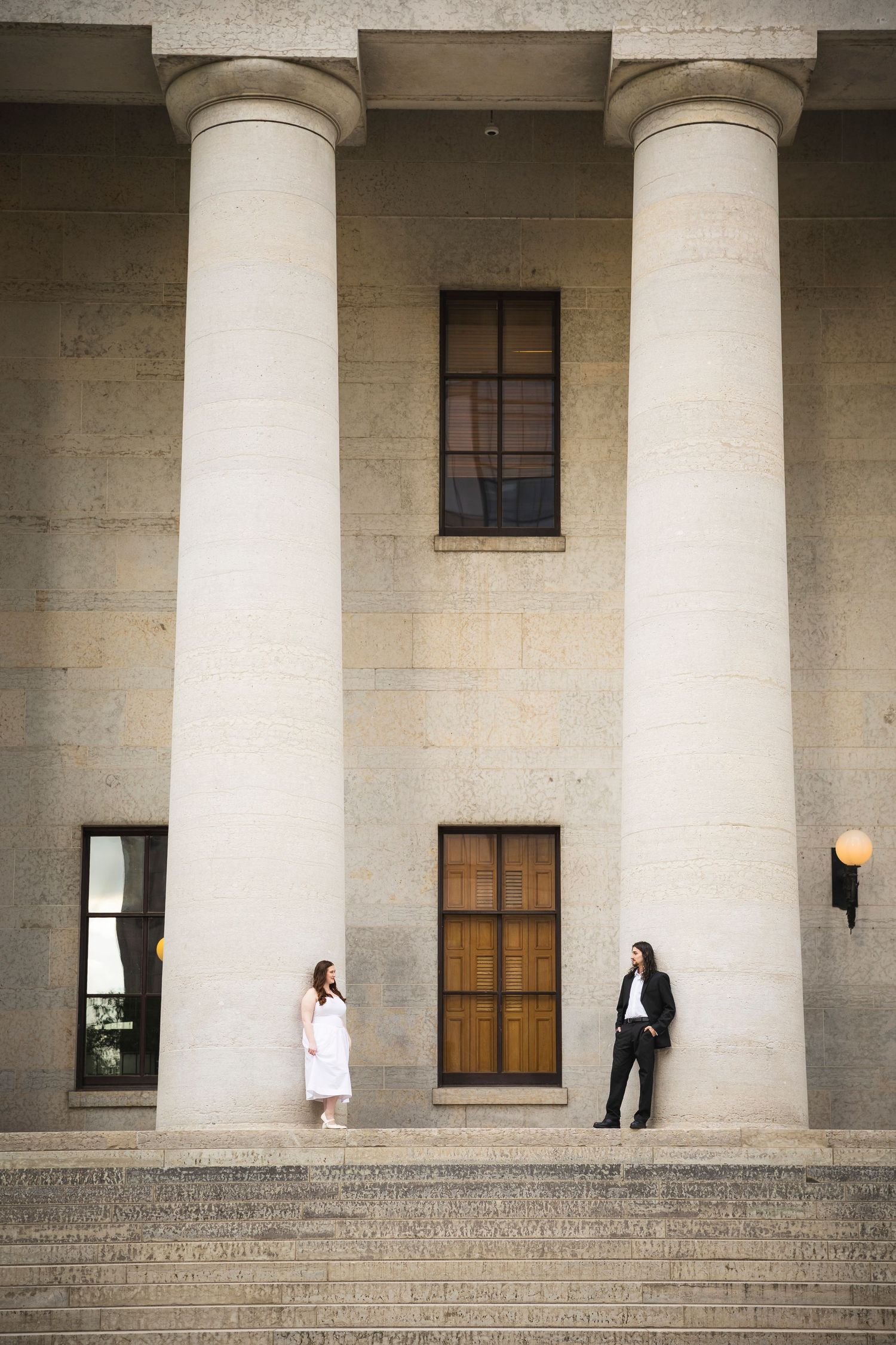 A couple poses romantically between elegant white columns in a classical building.