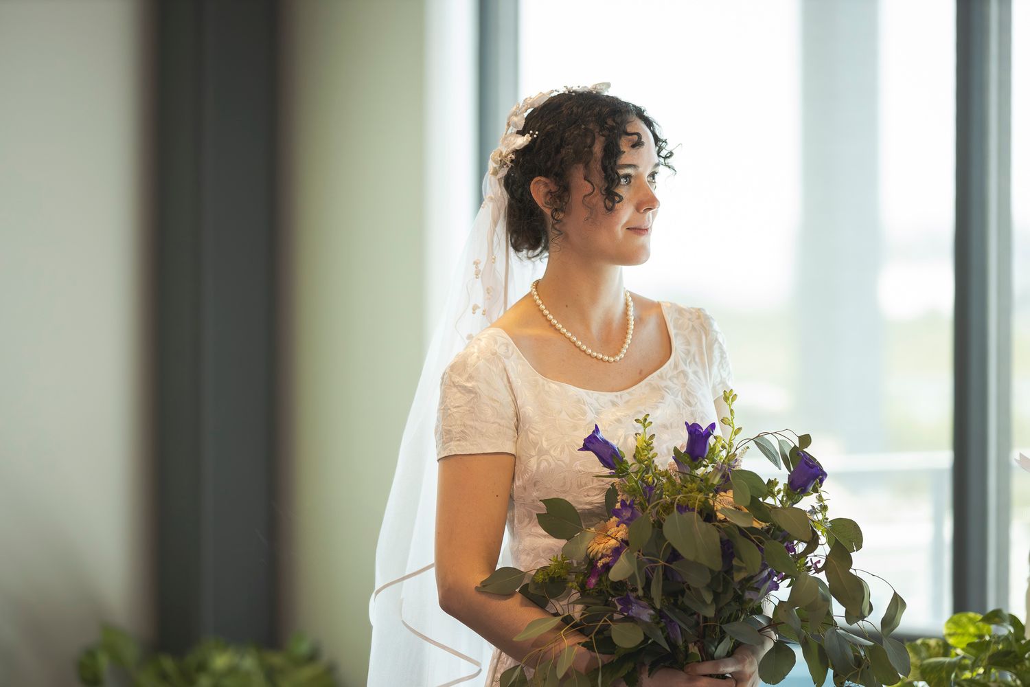 Bride holding a vibrant purple and green wedding bouquet near sunlit windows.