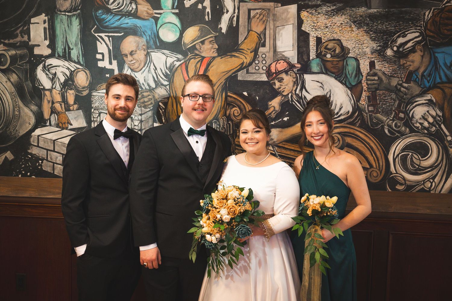 Wedding party poses in formal attire against decorative mural backdrop in historic venue.