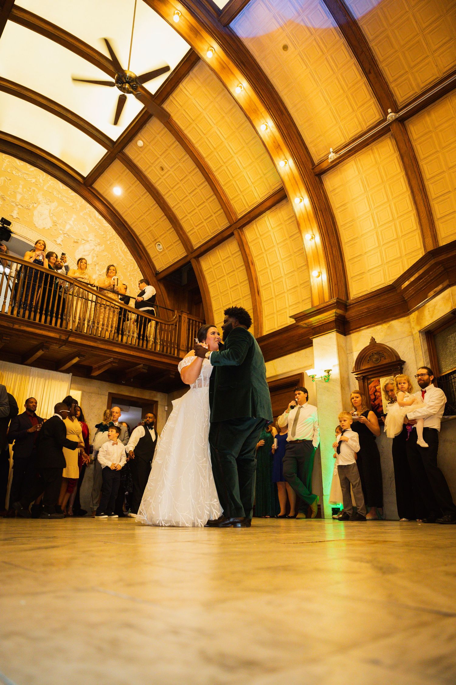 Wedding guests watch a couple's first dance in a grand ballroom with dramatic arched ceiling and ambient lighting.