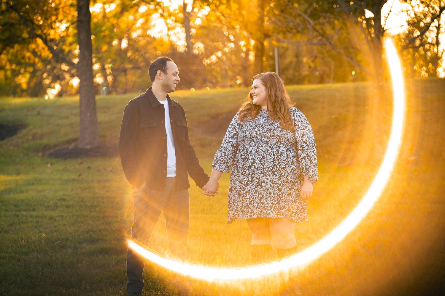 Dramatic lens flare creates a glowing arc around a couple during golden hour in a park setting.
