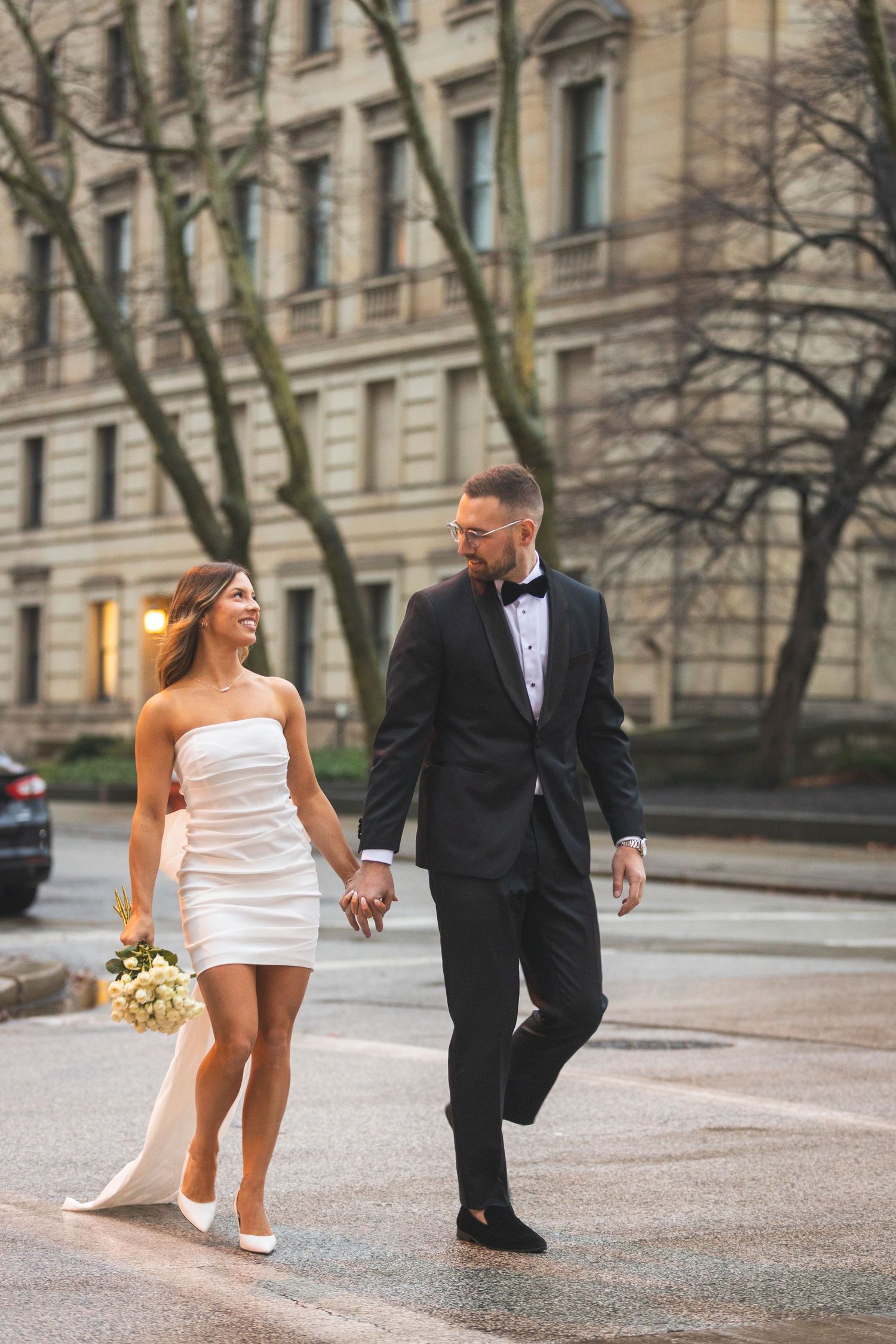 A couple in wedding attire walking hand in hand on a rainy city street.