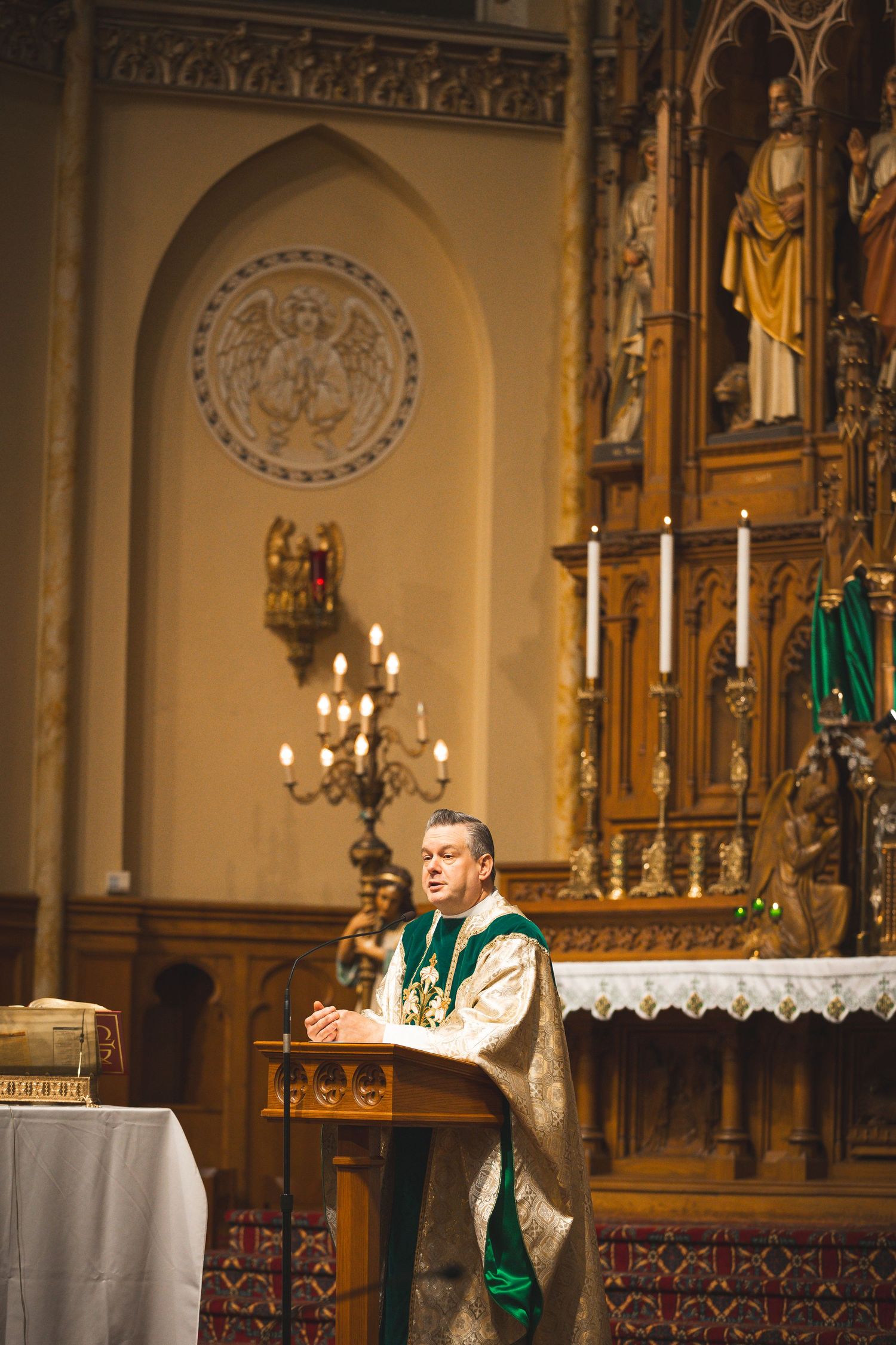 Priest conducting a religious ceremony at an ornate church altar with candles and green vestments.