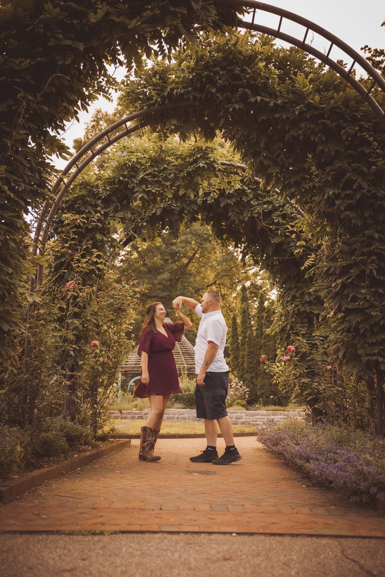 A couple stands beneath a curved garden arbor surrounded by greenery on a brick path.