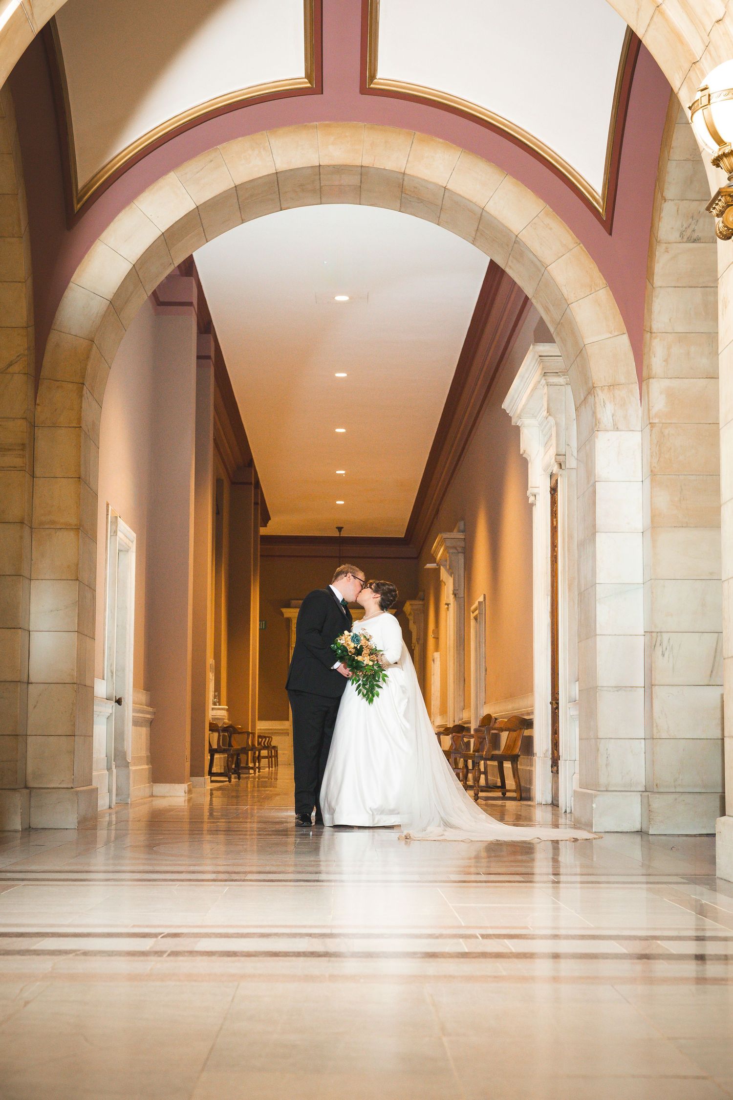 A wedding couple poses in an elegant arched hallway with stone columns and marble floors.