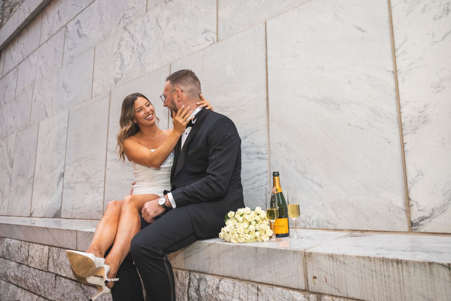 A candid moment between newlyweds sitting on stone steps with champagne and white roses.