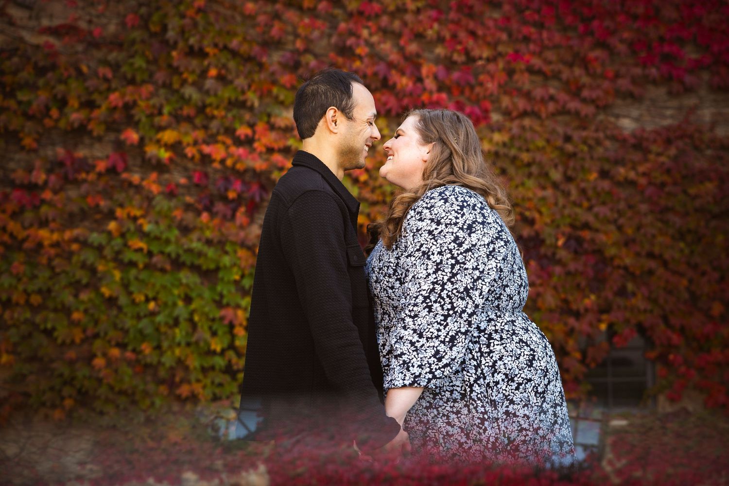 A couple poses against a wall of vibrant red and orange autumn ivy.