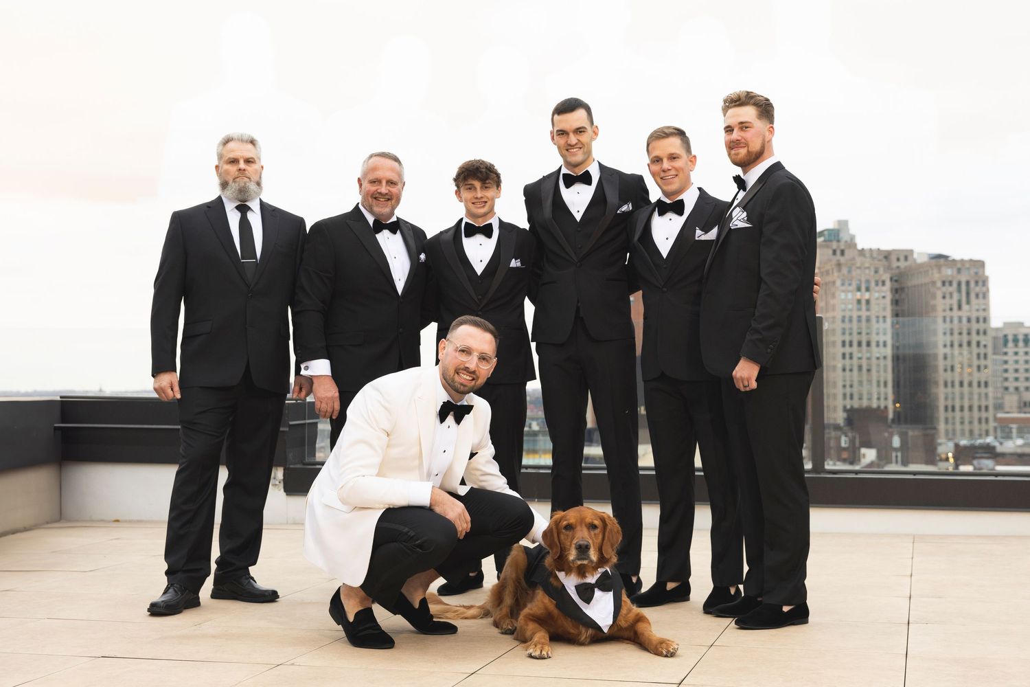 Wedding party in formal attire poses with dog on rooftop against urban backdrop.