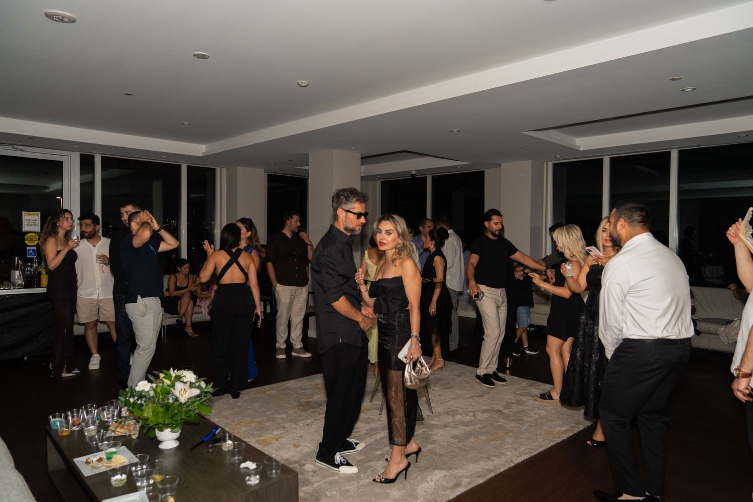 People mingling and dancing at a social event in a modern venue with white decor and floor-to-ceiling windows.
