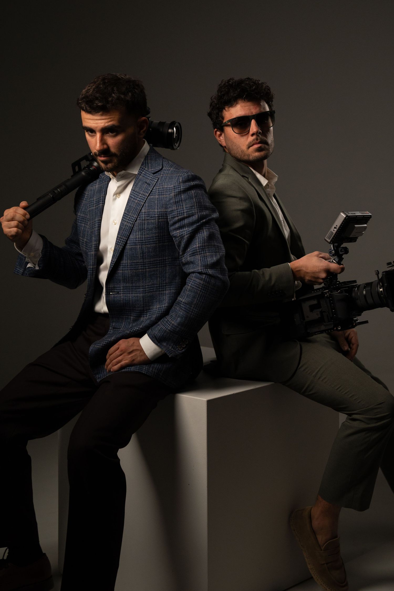 Two people in suits and sunglasses pose dramatically in a film noir style photoshoot against a dark background.