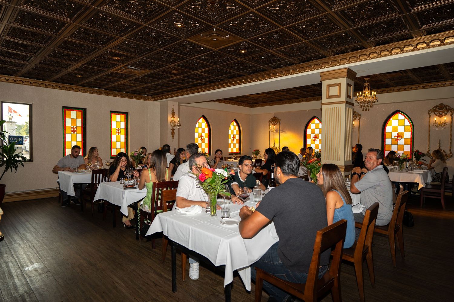 Elegant restaurant dining room with arched windows and ornate ceiling filled with guests at white-clothed tables.