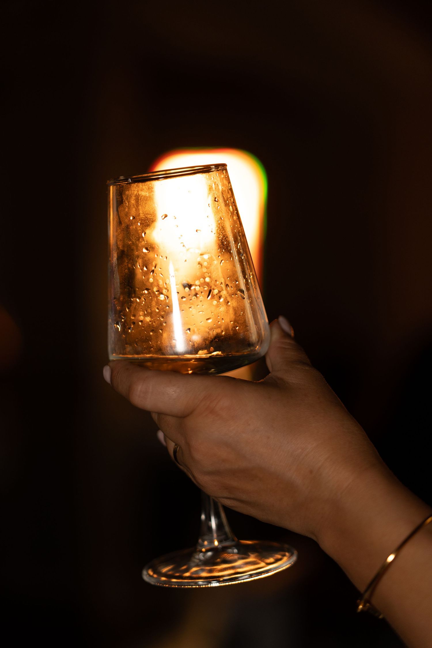 Hand holding a glowing wine glass illuminated by warm candlelight in dark setting.