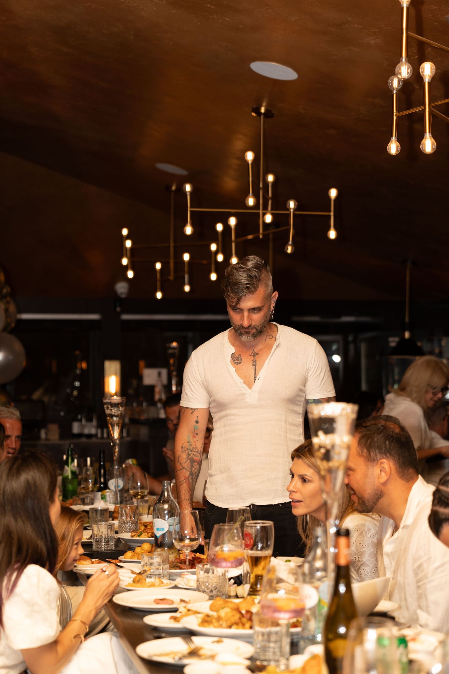 A dinner party scene with elegant hanging lights and guests gathered around a table filled with food and wine.